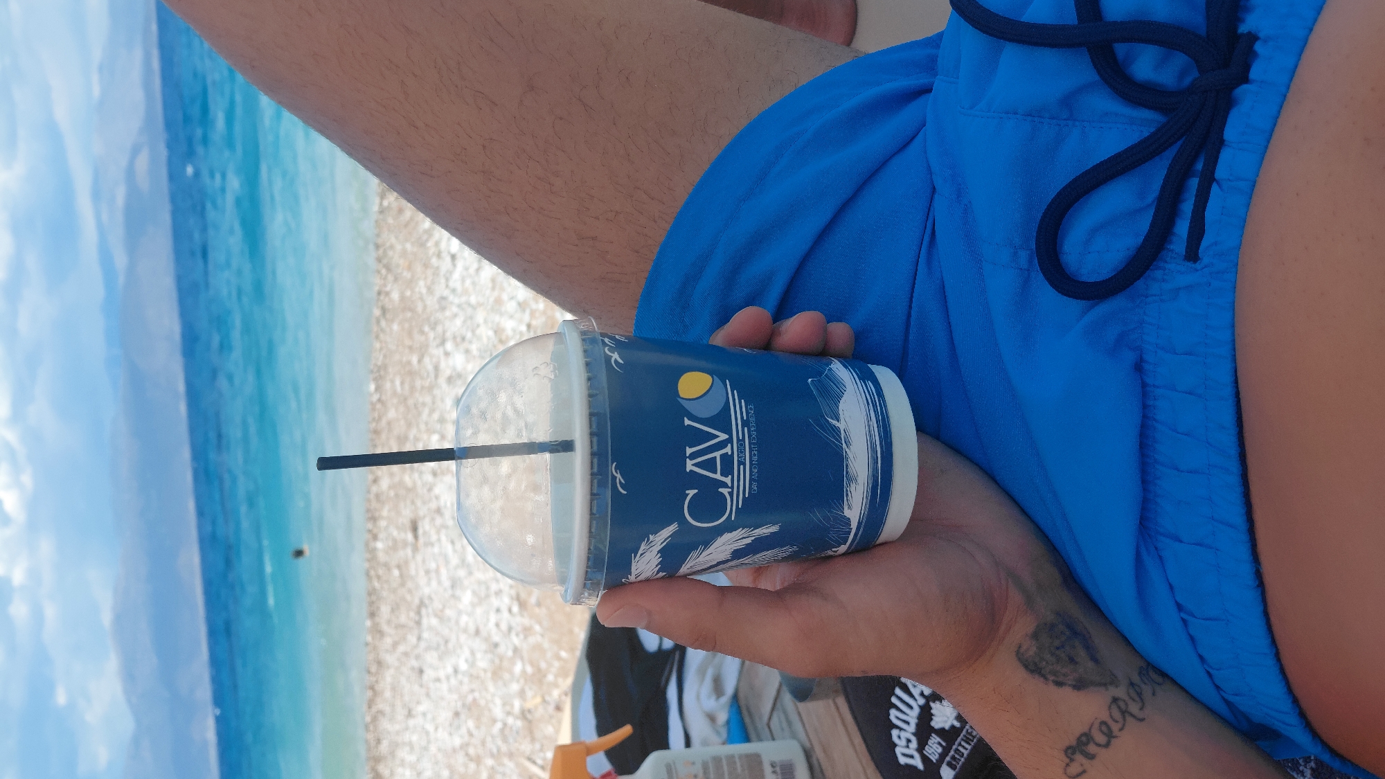 A person holds a blue disposable cup with a clear plastic lid and a black straw in their right hand. The cup has a white and blue design featuring palm trees, waves, and the text "CAVO" with "AIGIO" and "DAY AND NIGHT EXPERIENCE" below it. The person is wearing blue shorts. In the background, a beach with pebbles is visible, leading to a turquoise sea and distant mountains under a cloudy sky. The scene appears to be at a beach resort, likely in the Municipal Unit of Aegio, Greece, given the cup's branding.