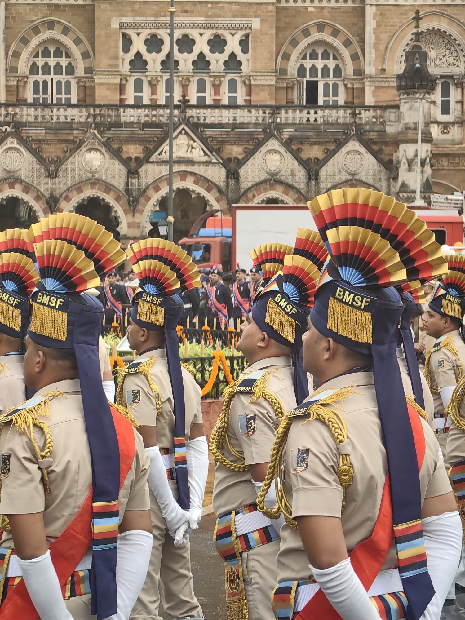 The image shows a group of people in uniform standing in front of a large, ornate, brown brick building. The building appears to be a historic landmark, possibly a government building or a train station, with arched windows and intricate architectural details. The people are all wearing similar tan uniforms with blue and red sashes. They have distinctive headdresses that are fan-shaped and colorful, with the letters "BMSF" visible on some of them. Their faces are not clearly visible, as they are either turned away or in profile. The overall scene suggests a formal event or parade. Based on the architectural style of the building, it is highly likely that the location is the Chhatrapati Shivaji Maharaj Terminus (formerly Victoria Terminus) in Mumbai, India, a UNESCO World Heritage Site known for its Victorian Gothic Revival architecture. The people appear to be part of an official procession or ceremony.