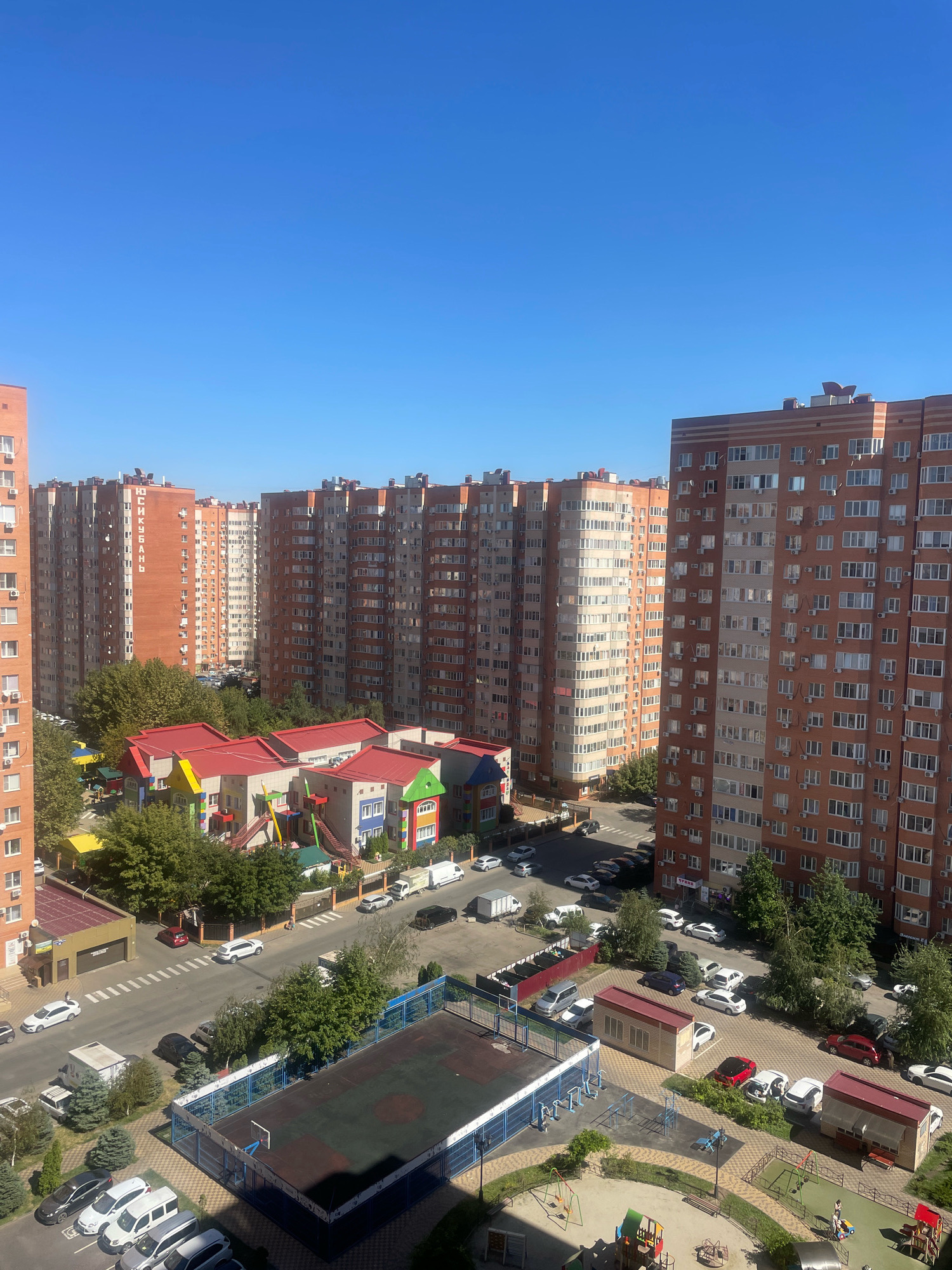 The image shows a residential area in Krasnodar, Russia, on a sunny day. The scene is dominated by several tall, brick apartment buildings. In the foreground, there are children's playgrounds and a sports court. A street runs between the apartment buildings and the playgrounds, with several cars parked or driving on it. There is also a small kindergarten or preschool building with a red roof. The sky is clear and blue. There are no identifiable people in the image.