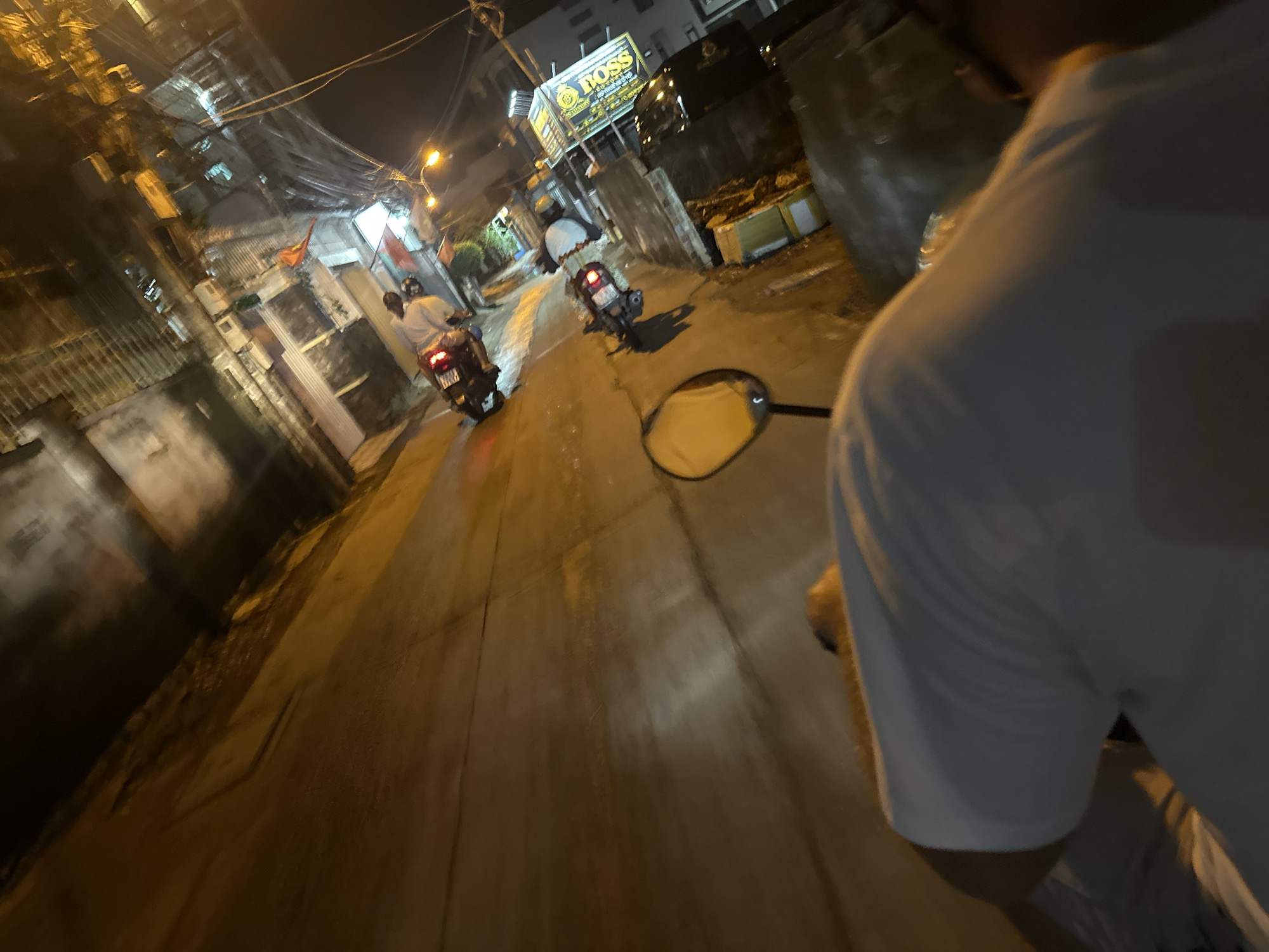 The image shows a nighttime street scene in Bac Nha Trang Ward, Vietnam. The photograph is taken from a low angle, likely from the perspective of someone riding a motorbike. Two other motorbikes are visible ahead, traveling in the same direction. Buildings line the street, with dense electrical wiring visible overhead. A sign for "BOSS" is illuminated. The street is wet, reflecting the ambient light. A motorcycle mirror is visible in the foreground, partially obscuring the view. The person taking the photo is wearing a white shirt.