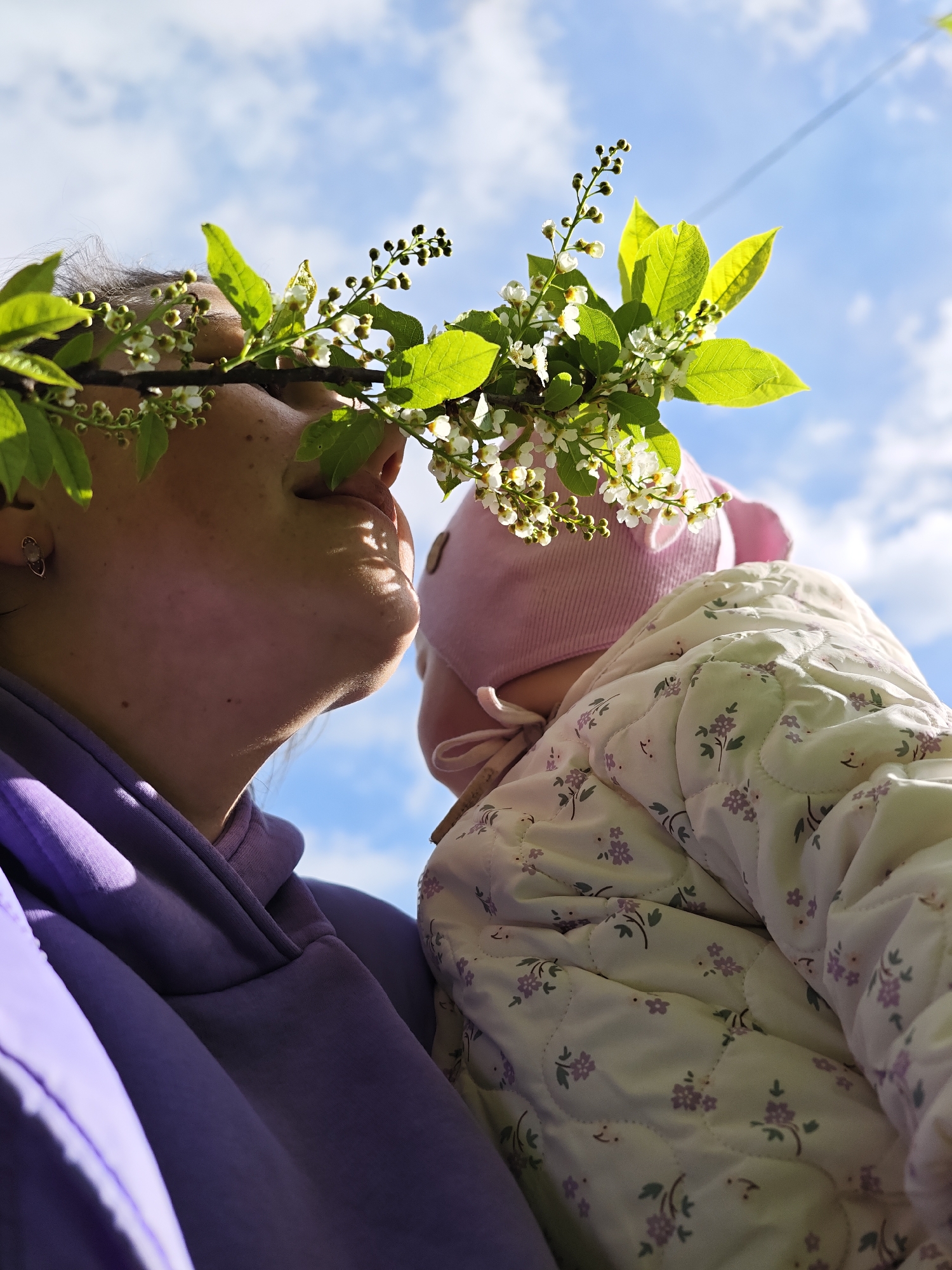 In Malyi Bukor, Russia, a low-angle shot shows a woman holding a baby outdoors against a backdrop of a partly cloudy sky. A blossoming branch with green leaves and small white flowers bisects the frame, partially obscuring their faces. The woman, wearing a purple hoodie, has her face tilted up with her eyes closed or nearly closed, suggesting she might be enjoying the sunlight or breeze. The baby, wearing a pink hat and a floral-patterned quilted jacket, is cradled in the woman's arms. The sunlight casts shadows and highlights on their faces and clothing, accentuating the texture of the fabric and the natural beauty of the foliage.