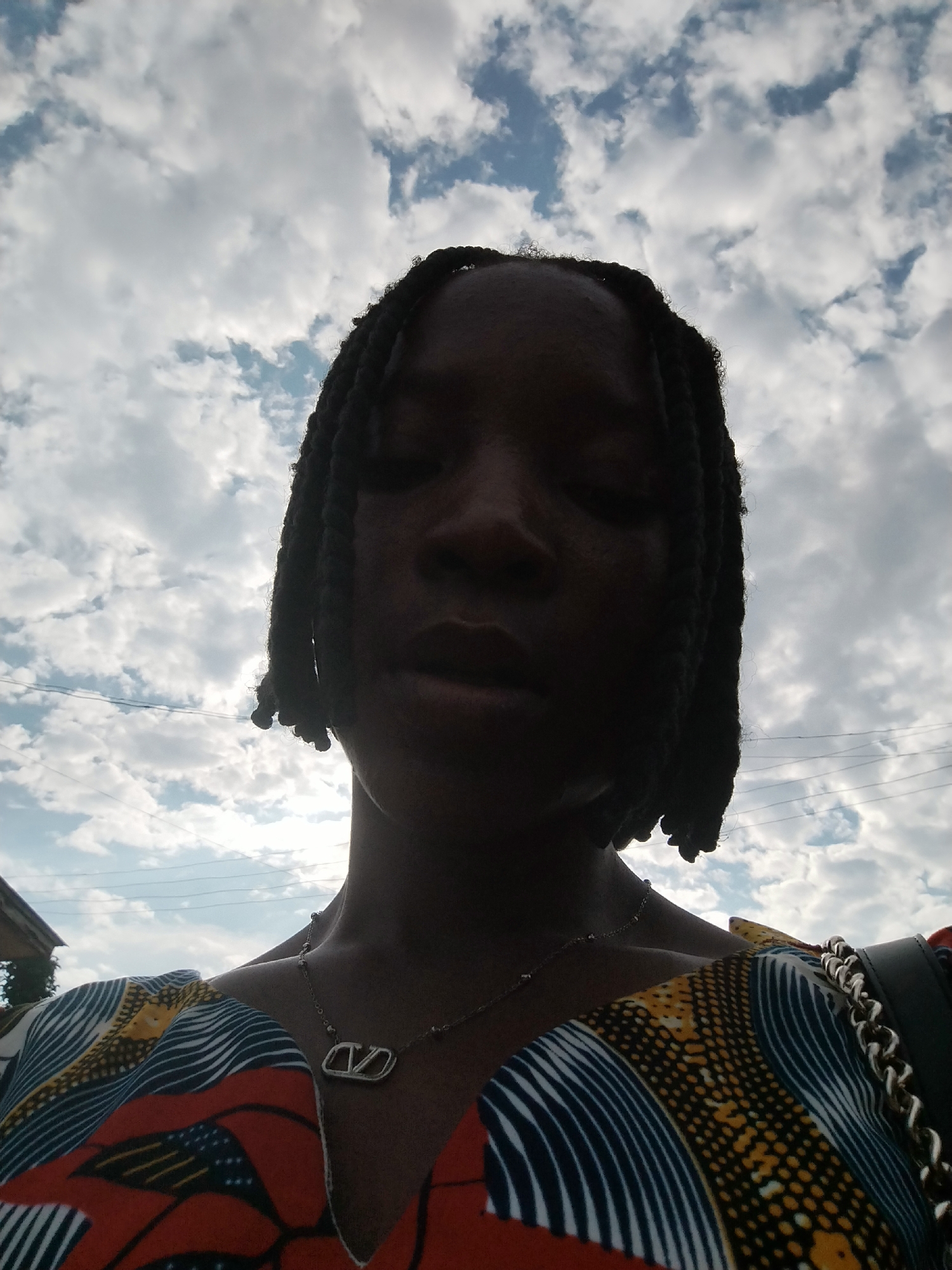 A close-up, low-angle selfie taken outdoors shows a person's head and shoulders against a cloudy sky. The person has dark skin and braided hair, with their eyes closed. They are wearing a brightly patterned top with a V-neck and a silver necklace with a pendant. A portion of a black bag with a chain strap is visible on the right side of the frame. The background consists of a cloudy sky with patches of blue. There are power lines visible against the sky and a hint of a building on the far left. The image appears to have been taken in Uselu, Nigeria, based on the context provided.