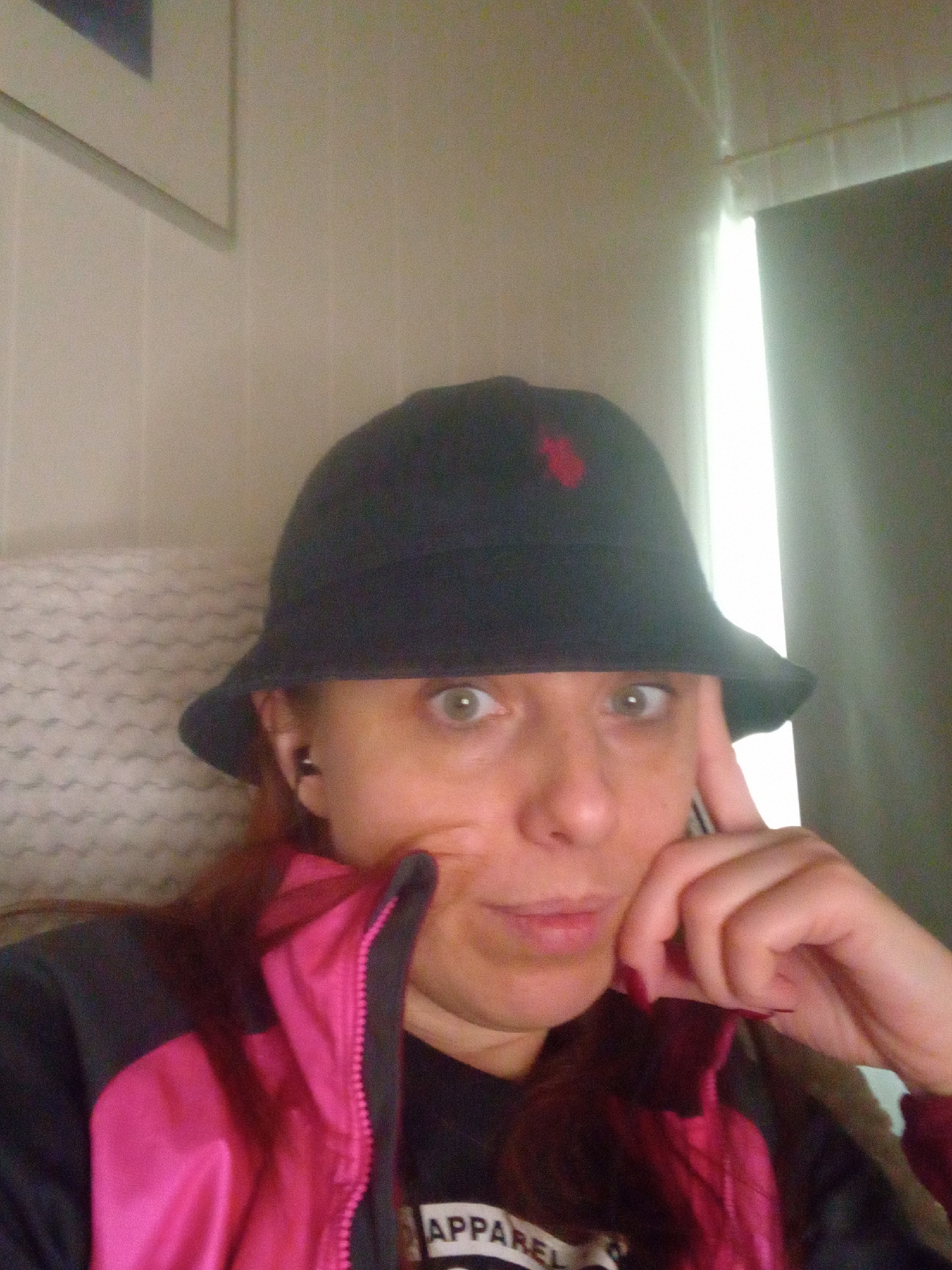 An adult individual is captured in a head-and-shoulders portrait shot within an indoor environment at an unknown location in Unknown, Unknown. The subject, appearing to be female, wears a black bucket hat with a small red embroidered emblem on the front, resembling a stylized horse and rider. Her eyes are wide and light in color, staring directly at the camera. Her hair, visible beneath the hat and around her shoulders, is reddish-brown. She is wearing a black and fuchsia (bright pink) zippered jacket, with the fuchsia visible on the lining and as a design element on the exterior. A black shirt with partially visible white text, reading "APPAREL," is worn underneath. A small black earbud is visible in her left ear. Her left hand is raised, with fingers resting near her chin and mouth. The background on the left features a light-colored wall with a framed picture. On the right, a window with vertical blinds or curtains is present, showing a bright vertical strip of light, indicating an external light source. A light-colored, patterned fabric, possibly a blanket or cushion, is visible behind the subject's left side. The image exhibits a slightly grainy texture.