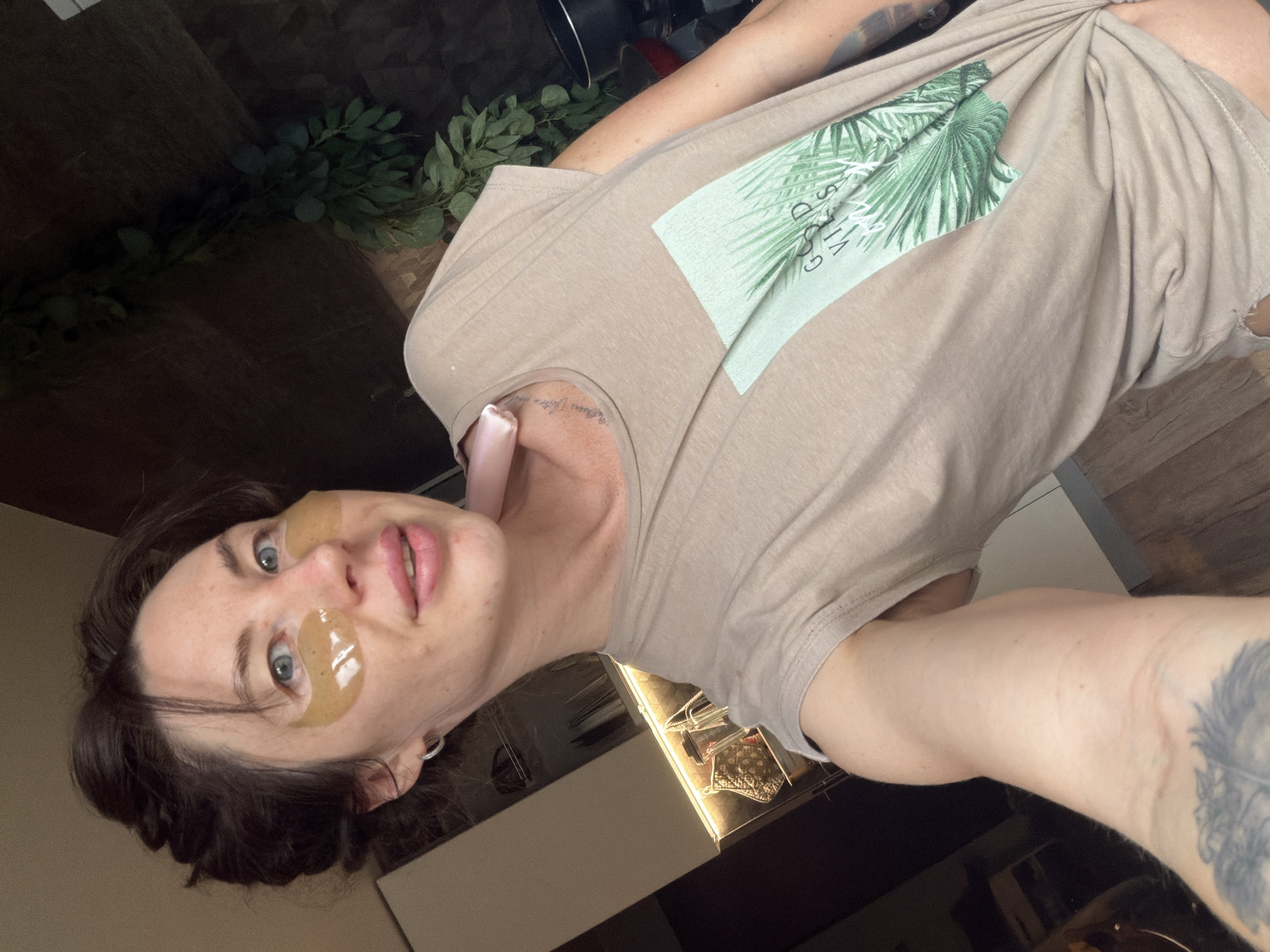 A medium shot shows a woman from the shoulders up, looking directly at the camera. She has dark hair pulled back, blue eyes, and is wearing under-eye patches. A pink device is visible near her neck. She is wearing a beige t-shirt with a palm leaf design and the words "GOOD VIBES" printed on it. The background appears to be a kitchen with cabinets and a hanging plant. The image does not contain information about the city of Perm, Russia.