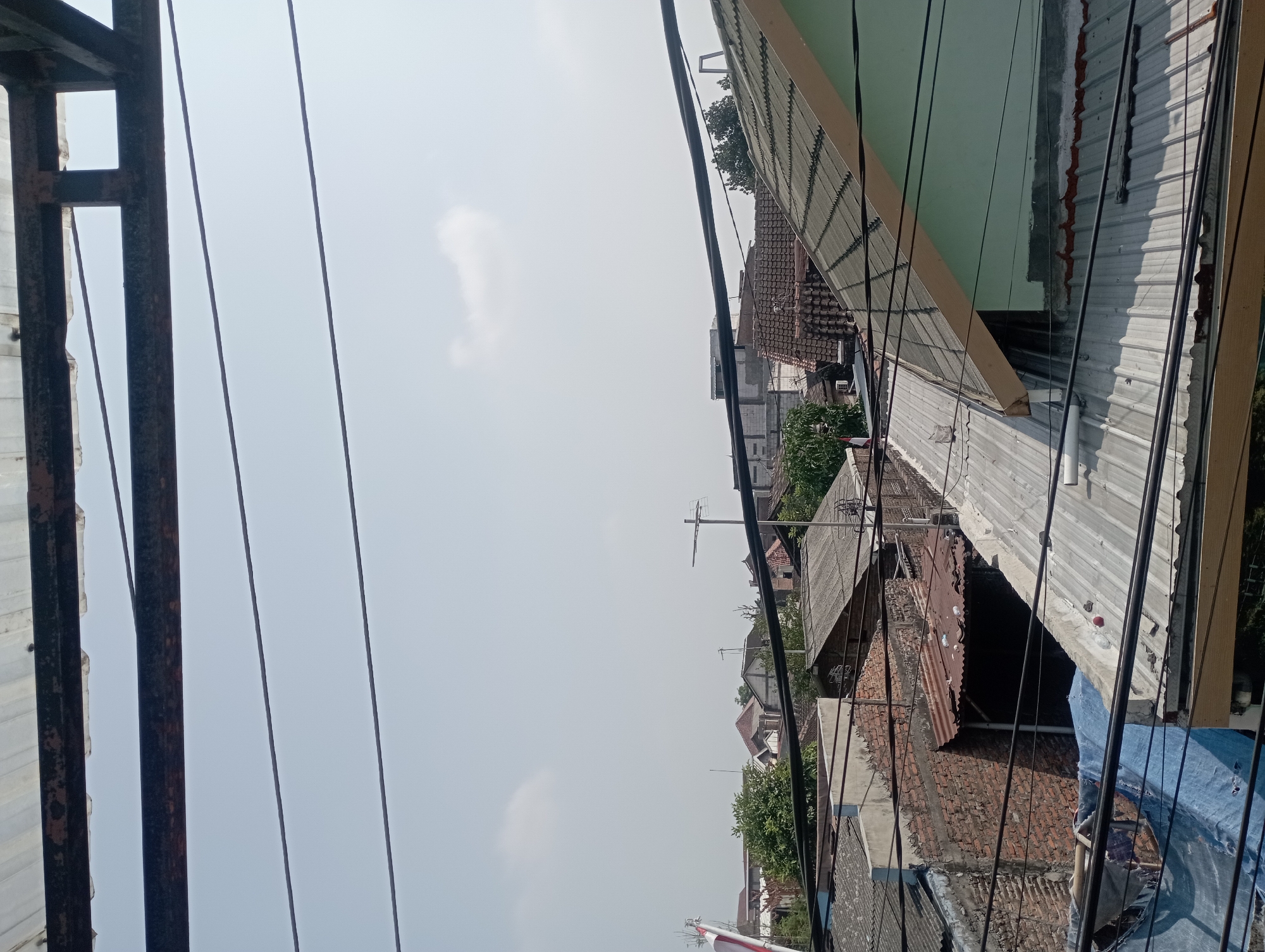 The image captures an urban scene in the city of Surakarta, Indonesia, viewed from a slightly elevated perspective. Dominating the foreground are several black electrical wires stretched across the frame, some appearing taut, others slightly sagging. Below the wires, a jumble of rooftops is visible. The roofs are constructed from various materials, including corrugated metal, tiles, and what appears to be some form of tarpaulin or plastic sheeting, one of which is a bright blue.
The buildings themselves exhibit a range of architectural styles and states of repair. Some walls are made of exposed brick, while others are plastered. There are visible signs of weathering and age on many of the structures. A light green painted wall of a building is prominent on the right side of the frame. Further into the background, more houses and buildings can be seen, interspersed with trees and vegetation. A flagpole with an Indonesian flag is partially visible on the left side of the image. The sky above is a pale, hazy blue with a few faint clouds. The overall impression is of a densely populated urban area, likely a residential neighborhood, under daylight conditions. There are no people clearly visible in the image.