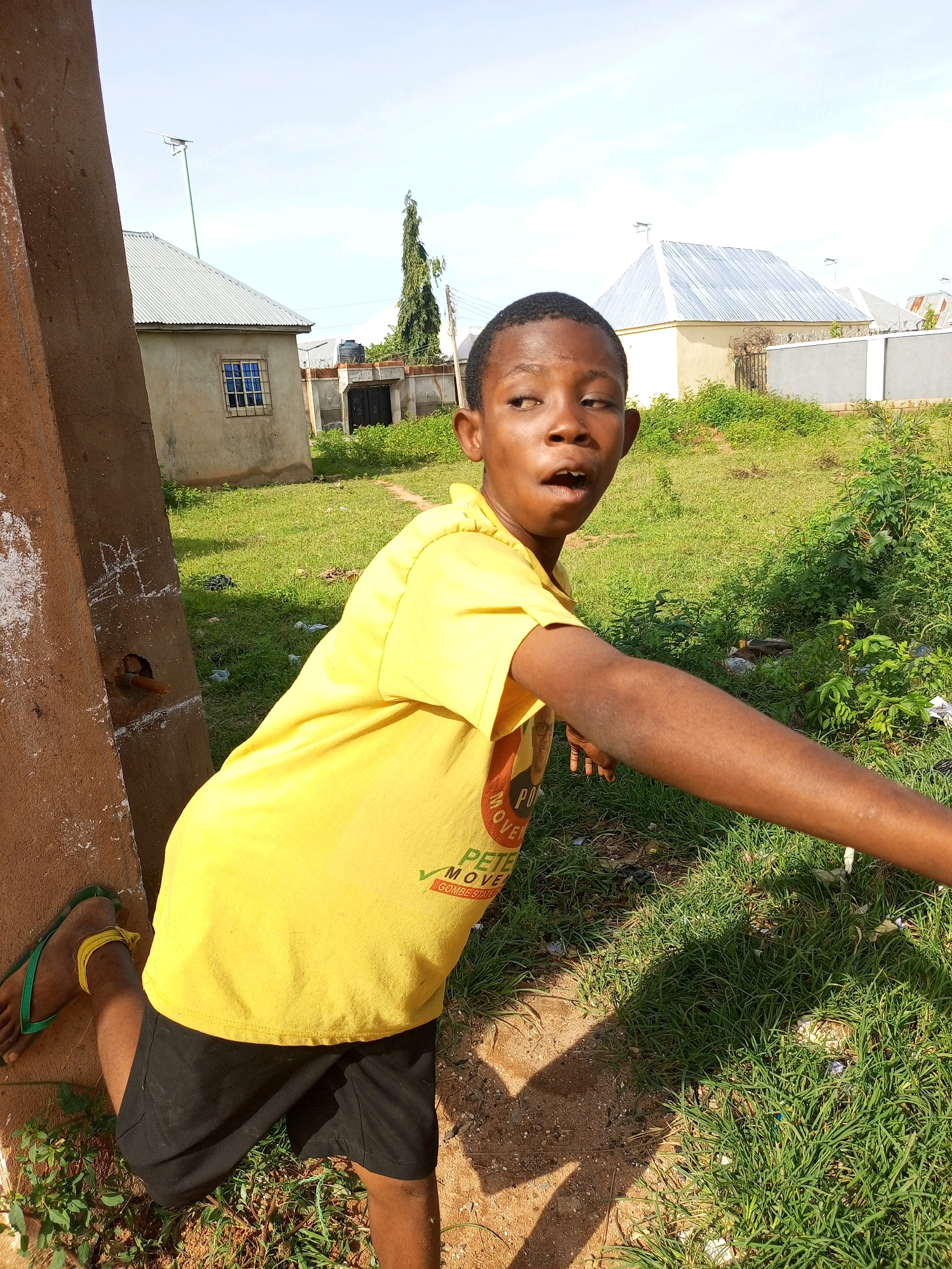 The image shows a young boy in Tempure, Nigeria, standing in an outdoor setting. He is wearing a bright yellow t-shirt and black shorts, with green flip-flops on his left foot. The boy is looking to his right with a surprised or excited expression, his mouth slightly open. He is leaning on a thick, brown post, with his right arm extended outwards. Behind him, the scene includes a grassy area with some scattered debris, a dirt path, and buildings with corrugated metal roofs. In the background, there are trees and a clear sky, suggesting it is daytime with fair weather. The boy's t-shirt has some visible text, including "MOVE" and "PETE", possibly related to a campaign or event. The overall impression is a candid shot capturing a moment of expression from the boy in his environment.