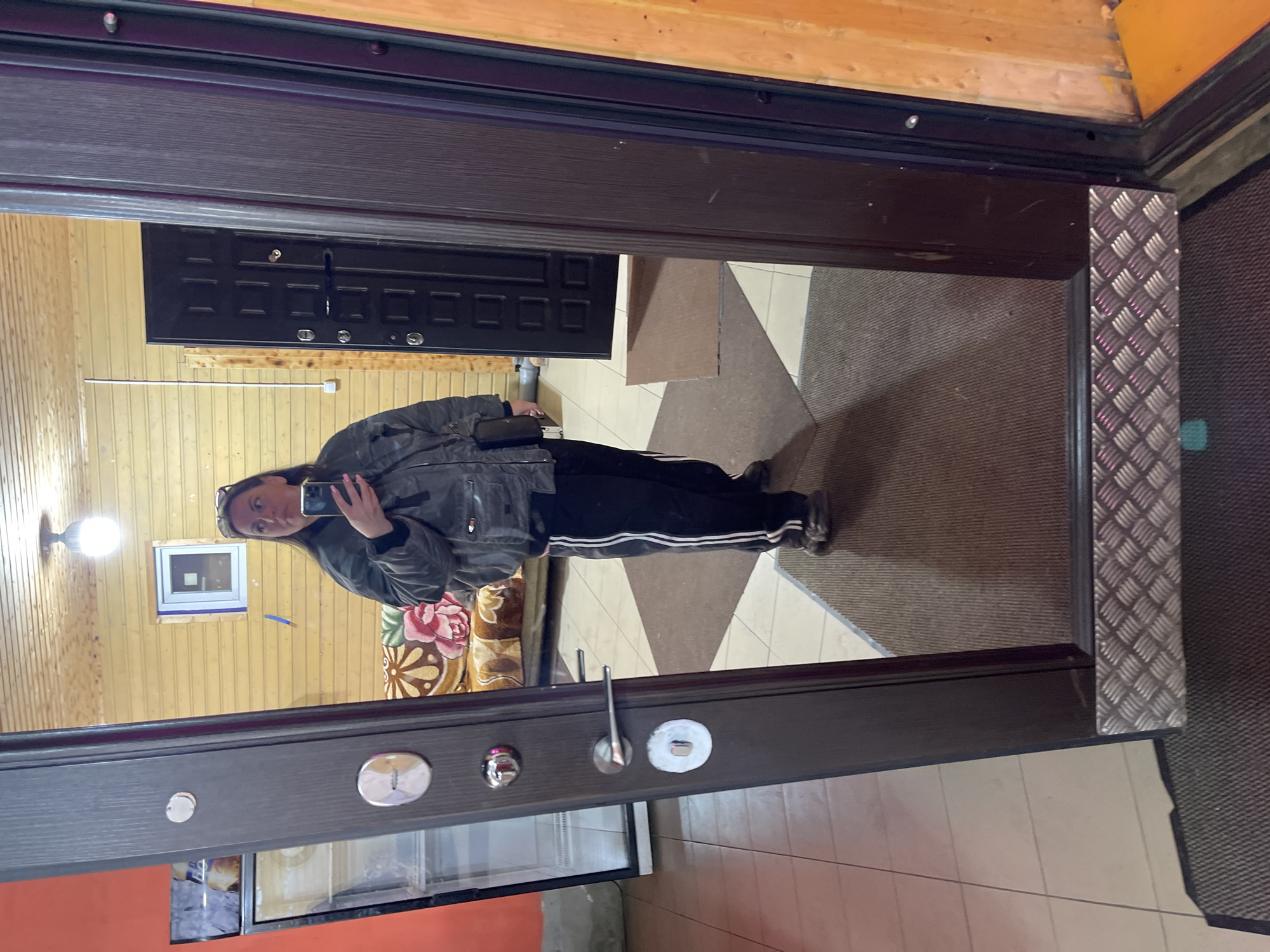 The image shows a person reflected in a mirror, presumably in Картмазово, Russia, although this location information is speculative. The person is holding a phone and taking a photo of themselves in the mirror. The person is wearing dark pants with white stripes and a gray jacket.
The mirror is part of a door. The reflection in the mirror shows a room with wooden walls, a window, and a black door. The floor in front of the door appears to be tiled. The background visible through the door reflection includes part of a sofa with a floral pattern.