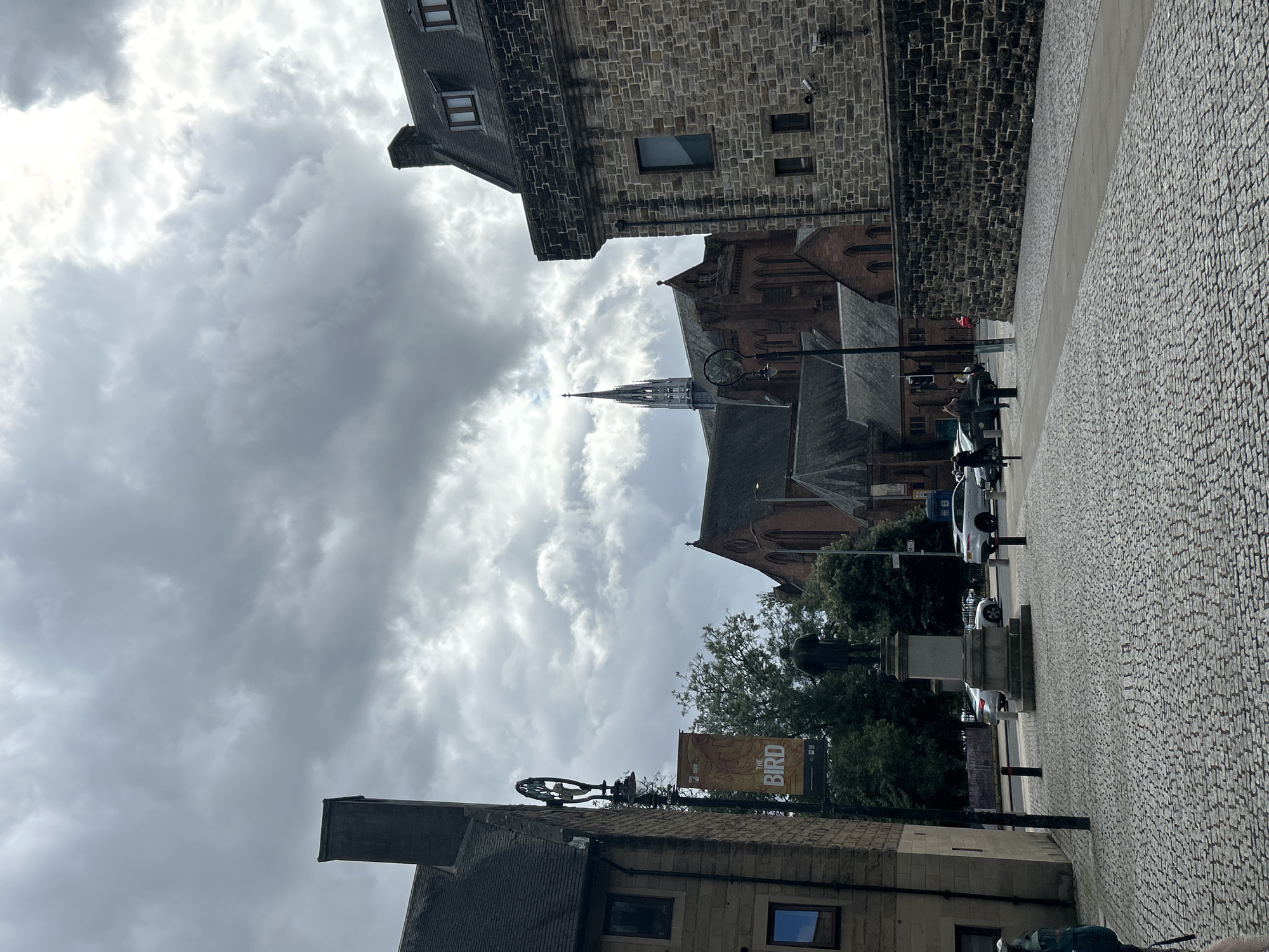 The image shows a wide cobblestone plaza in front of stone buildings and a red brick church with a spire in Glasgow, United Kingdom. A statue of a man stands in the center of the plaza. A banner with the word "THE BIRD" is hanging from a lamppost. Several cars and pedestrians are visible in the background. The sky is overcast with dramatic clouds.