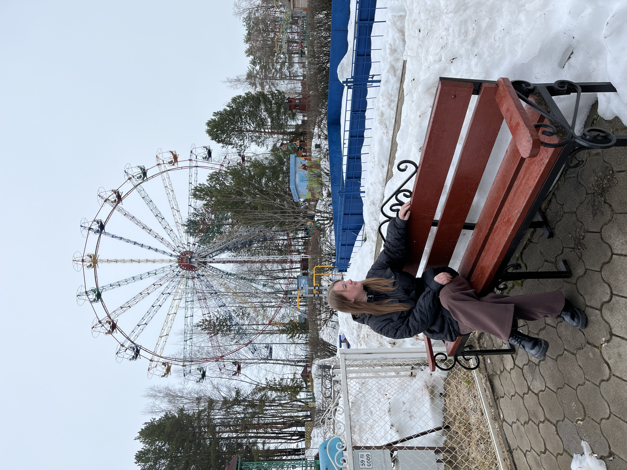 A young person is sitting on a red bench with decorative black metal armrests. The person is wearing a black jacket, brown pants, and black shoes. In the background is a Ferris wheel and bare trees. Some snow is visible on the ground. The photograph appears to be taken in an amusement park in Chaikovsky, Russia.