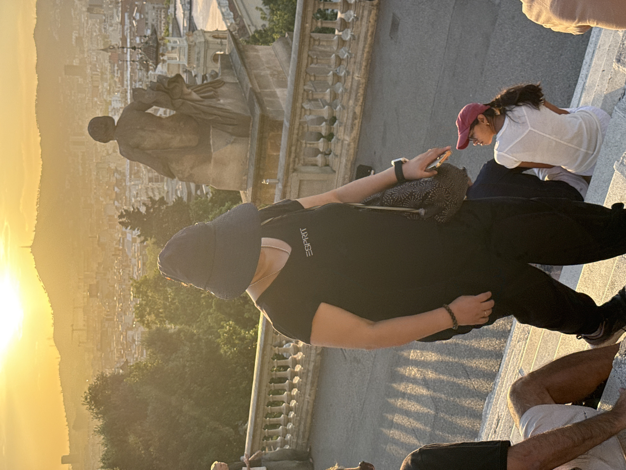 This image captures a late afternoon scene with the sun setting over Barcelona, Spain, casting a warm glow and long shadows across a city overlook. In the foreground, a person wearing a black t-shirt with the "Esprit" logo and a dark wide-brimmed hat stands on a stone staircase. They are looking towards the right side of the frame. To their right, a young woman with dark hair pulled back in a ponytail, wearing a white t-shirt and a maroon baseball cap, is seated on the steps, looking upwards and to the left. The background reveals a panoramic view of Barcelona, with buildings stretching towards a distant, hazy mountain range. Prominent in the middle ground is a stone balustrade and a large stone statue on a pedestal. The overall impression is of a tourist gathering at a viewpoint, enjoying the sunset and the cityscape.