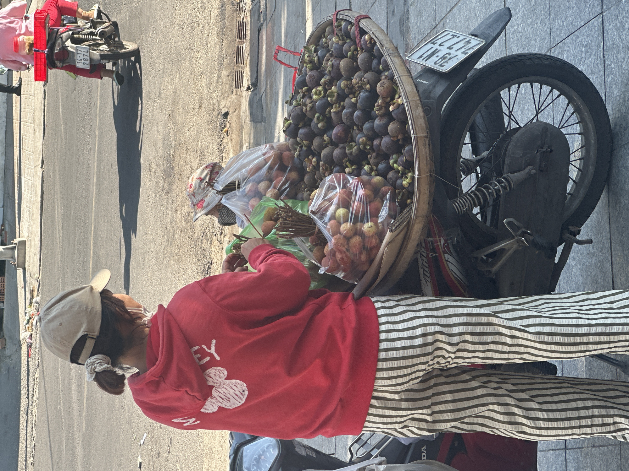 In Bac Nha Trang Ward, Vietnam, a person with a cap and a red hoodie stands next to a motorcycle. The motorcycle has a basket attached to it filled with mangosteens and lychees. The license plate on the motorcycle reads "LZ-72 LN-62." The person is sorting through the lychees, which are in plastic bags. The background shows a city street with a sidewalk.