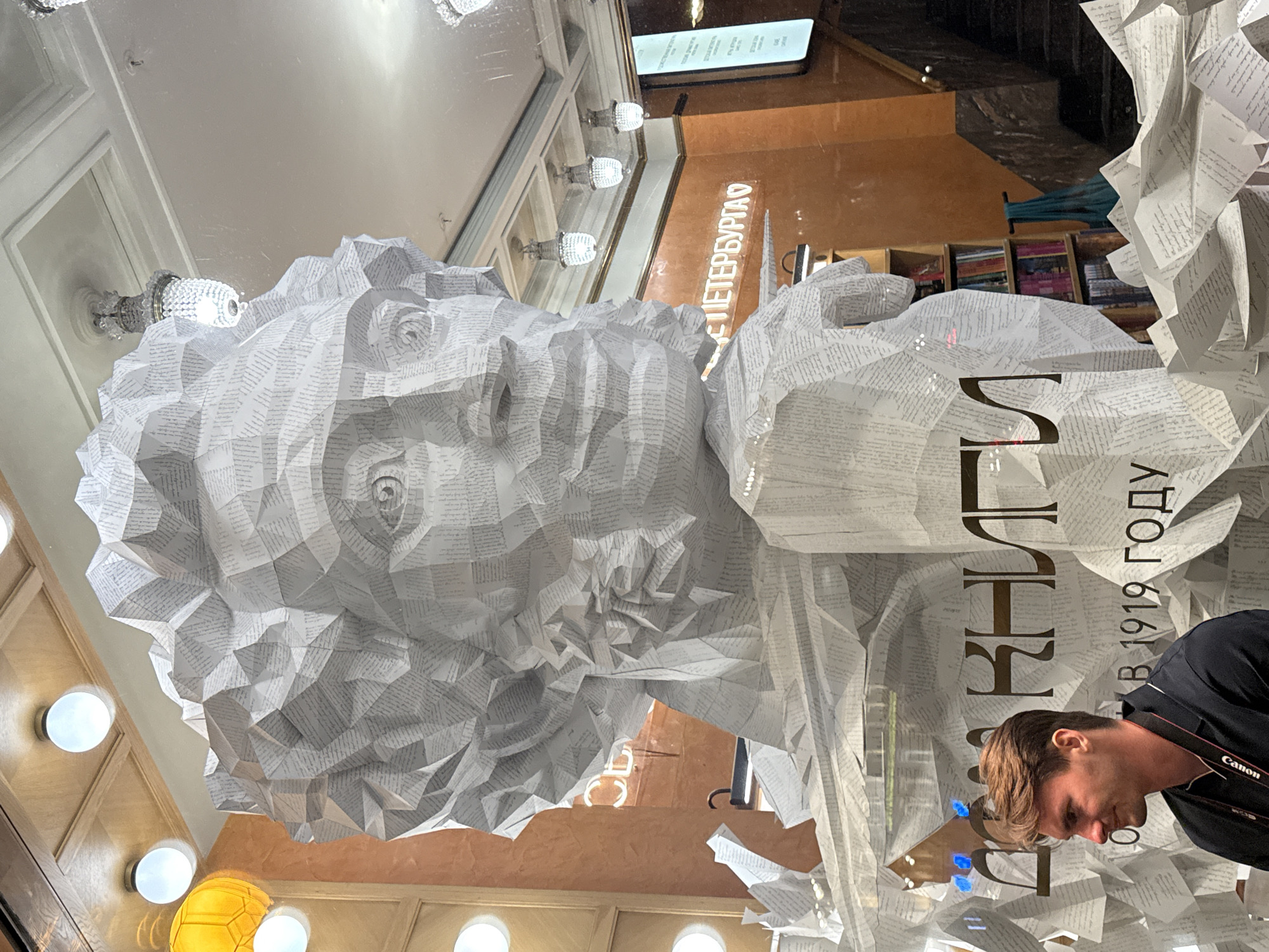 The image shows a large sculpture of a human head and shoulders, made entirely of folded paper that appears to be from books. The sculpture is highly detailed, with faceted surfaces creating the illusion of a face and hair. Behind the sculpture, text is visible on the wall and on more paper structures. The text is in Russian, with the words "ДОМ КНИГИ" (Dom Knigi) and "В 1919 ГОДУ" (in 1919) clearly legible. Another sign reads "ПЕТЕРБУРГ" (Petersburg). A young man with brown hair, wearing a dark collared shirt and a camera strap, is visible in the lower left corner, looking to the left and slightly down. The environment appears to be an interior space, likely a shop or exhibition, with chandeliers and lighting fixtures visible in the background. The overall impression is of a creative display within a bookstore, possibly celebrating the establishment's history.