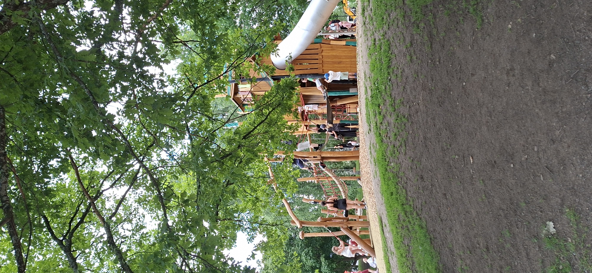 The image depicts a playground area at the Pikku-Vesijärvi park in Lahti, Finland. The playground features a large, multi-level wooden structure with various climbing elements, including ropes and nets, and a prominent silver slide. Several children and adults are visible interacting with the playground equipment. Some are climbing, while others are standing on platforms or descending the slide. The playground is surrounded by lush green trees, providing a shaded environment. The ground in the foreground is a mix of dirt and grass. In the background, more trees and some buildings are visible, indicating an urban park setting. A yellow construction vehicle is partially visible in the far right background.