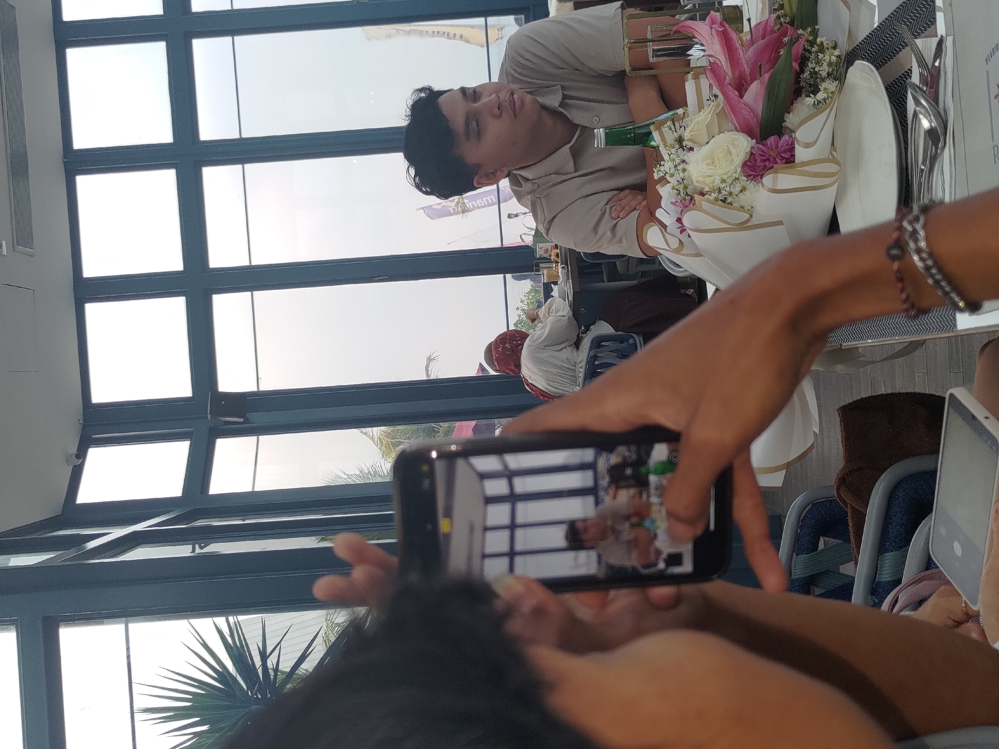 In Kosambi Barat, Indonesia, at a restaurant with large windows overlooking a bright, overcast sky, a person is taking a photo of another person sitting at a table. The person taking the photo is holding a smartphone in the foreground, with their arm and shoulder visible. The smartphone screen displays a reflection of a man seated at a table.
The man in the reflection, and also visible in the main image, is wearing a light brown collared shirt and has his arms crossed. He is looking to his right. In the background, a woman wearing a red hijab is seated at another table. A floral arrangement with white roses and pink lilies is on the table in front of the man being photographed. A green bottle and a glass are also on the table. The environment appears to be a dining establishment, possibly with an outdoor seating area given the presence of palm fronds visible through the windows.