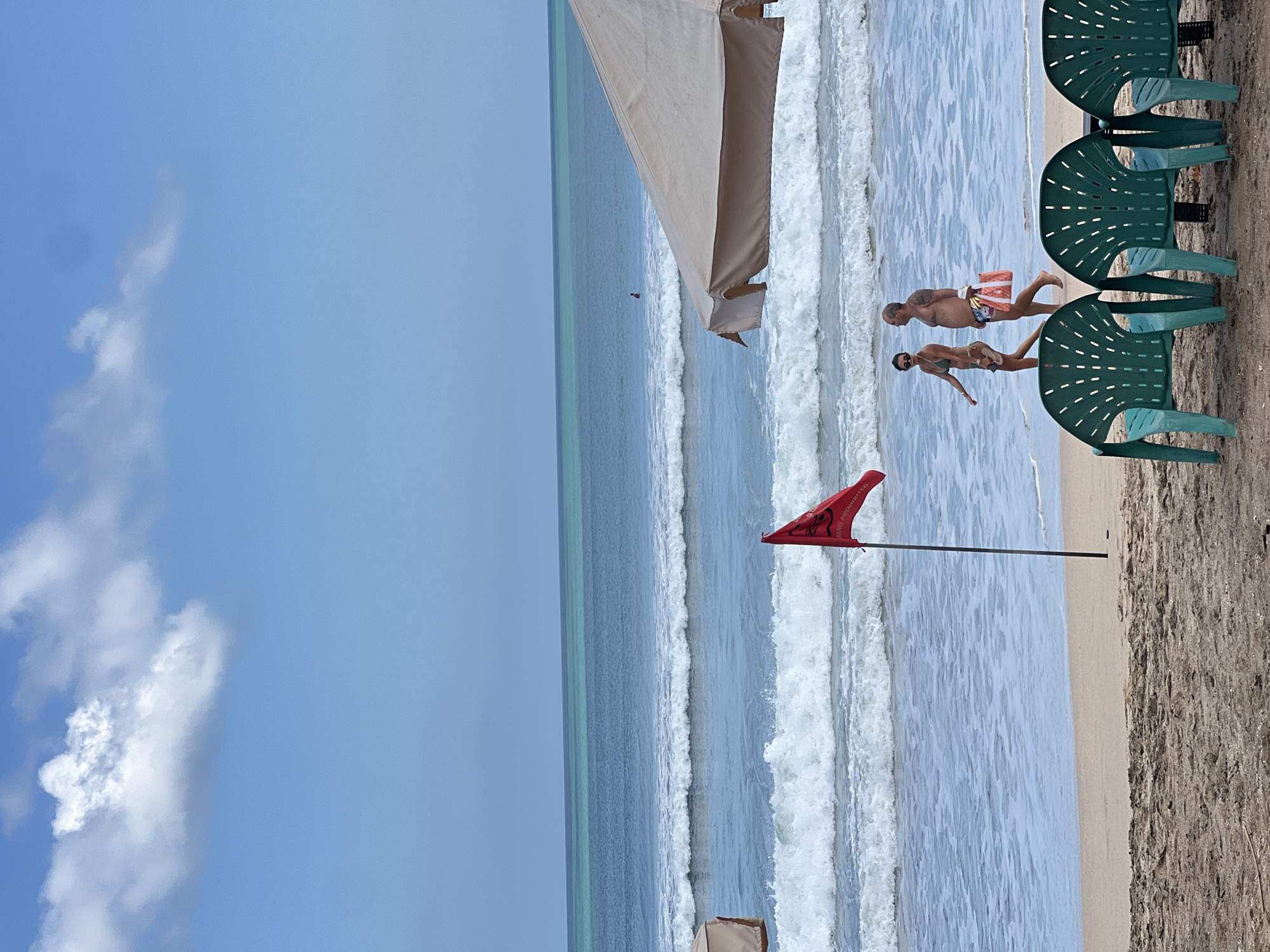 In Kuta, Indonesia, the image captures a serene beach scene under a bright, partly cloudy sky. The ocean stretches out in shades of turquoise and deep blue, with gentle waves rolling onto the sandy shore. A red flag, indicating swimming is prohibited, stands planted in the sand near the water's edge. In the background, a person is visible in the ocean, appearing to be a surfer or swimmer.
In the mid-ground, a couple walks along the beach. The woman, wearing a bikini, is on the left, and the man, in swim trunks, is on the right. He carries a bag. A large, beige umbrella provides shade in the foreground, partially obscuring the view of the ocean. In the immediate foreground, three green plastic chairs are arranged on the sand, suggesting a seating area for beachgoers. The overall impression is one of a calm and relaxing day at the beach.