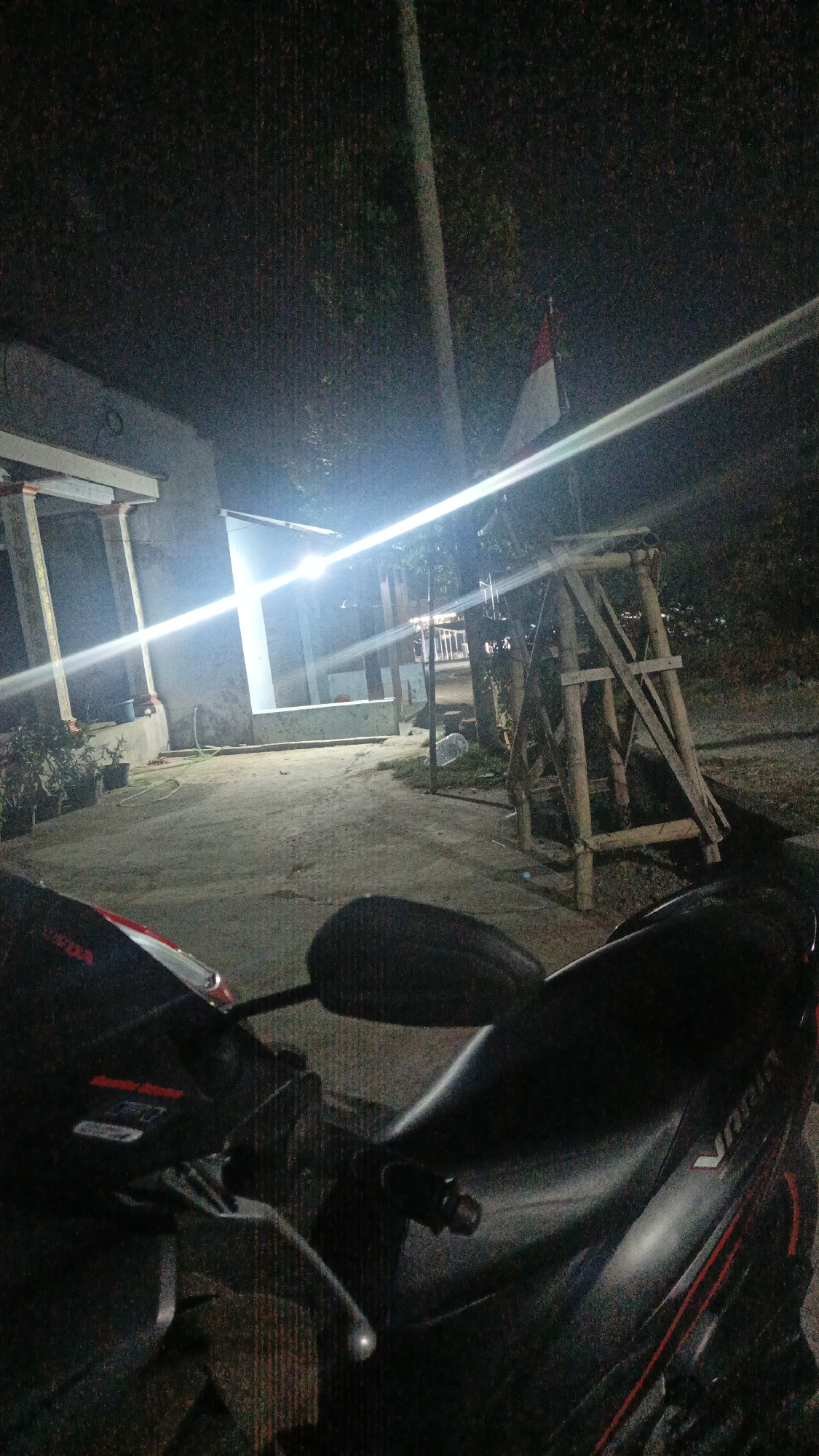 The image depicts a nighttime outdoor scene, likely in a residential or semi-rural area within Sambi, Indonesia. In the foreground, the handlebars and part of the seat of a dark-colored motorcycle are visible. The background shows a simple building with a porch and pillars, illuminated by a bright, horizontal light source, possibly a floodlight. To the right of the building, a wooden scaffolding structure is present. A flag, appearing to be the Indonesian flag due to its red and white colors, is attached to a pole behind the scaffolding. The ground is paved with concrete and there are some potted plants near the building. The overall lighting suggests it is dark outside, with artificial lights providing illumination.