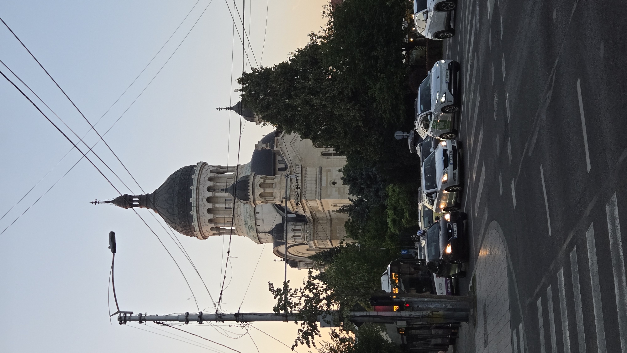 The image captures a street scene in Cluj-Napoca, Romania, featuring the Metropolitan Cathedral as the dominant background element. The cathedral is a large, ornate religious building with multiple domes and towers, constructed primarily of light-colored stone. In the foreground, a busy intersection is depicted with several cars stopped at a red traffic light, indicating a moment of pause in urban traffic. A pedestrian crossing is clearly visible, marked by white stripes on the asphalt.
Vehicles present include a black sedan with license plate CJ 19 SOY, a white station wagon with license plate CJ 07 IAL, and a white electric car with "Bolt" branding and license plate CJ 03 VPU. Other vehicles are partially visible in the traffic queue. A bus with the number 33 and destination "OREZISH" is also stopped at the intersection.
The environment is characterized by utility poles with numerous wires crisscrossing the sky, a common sight in urban areas. Trees line the street, some of which partially obscure the lower sections of the cathedral. The sky is a pale, hazy blue, suggesting either early morning or late afternoon light. A street light is visible on a pole to the left of the frame. No people are clearly discernible in the image.