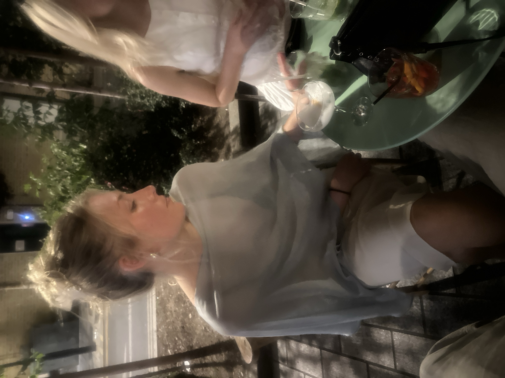 The image depicts two young women seated at an outdoor table in what appears to be London, United Kingdom. One woman, with long blonde hair, is wearing a white top and sitting across the table from the other. The second woman, with blonde hair pulled back into a bun, is wearing a light blue sheer top and a white skirt or dress. On the table, there are two cocktails - one clear with a white foam or garnish and another amber-colored drink with fruit. A black purse is also on the table. The background shows a blurred cityscape at dusk or night, with streetlights and buildings visible. The scene appears relaxed, suggesting the women are enjoying a social outing.