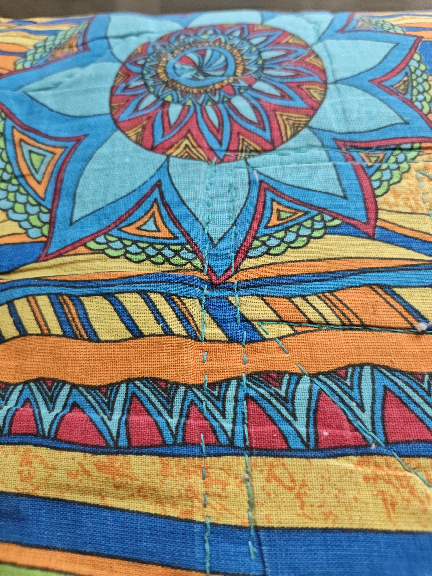 The image is a close-up shot of a colorful textile with a geometric and floral pattern. The dominant colors are bright blue, orange, yellow, red, and green. The fabric appears to be slightly textured, with visible stitching in a teal thread running vertically down the center and horizontally in some areas, suggesting it might be a quilted item or a decorative fabric panel. The pattern features repeating motifs that resemble stylized flowers or mandalas, surrounded by angular shapes and striped sections. The lighting is bright and even, highlighting the vibrancy of the colors and the details of the fabric's weave and design. There is no indication of a specific location in Shcherbinka, Russia, as the image focuses solely on the textile itself.