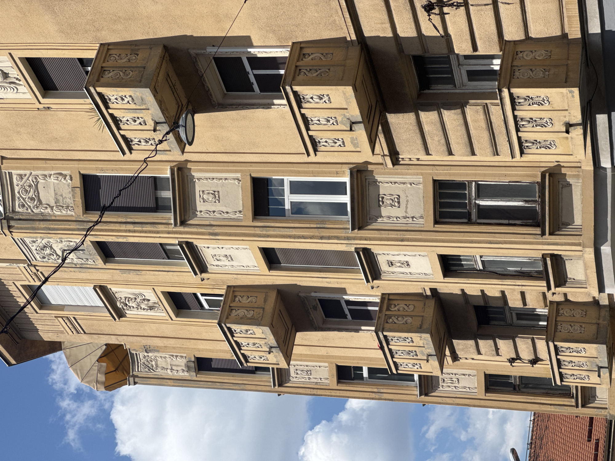 The image shows an exterior view of a multi-story building in Belgrade, Serbia. The building features a light brown facade with architectural details, including decorative relief panels and balconies with ornate railings. The balconies are cantilevered and appear to be made of stone or concrete. Several windows are visible, some with dark-colored shutters and others with white frames. Overhead, electrical wires run across the facade. In the upper left corner, a portion of a retractable awning is visible. The sky is blue with white clouds. The overall impression is of an older, well-maintained urban building with classical architectural influences.