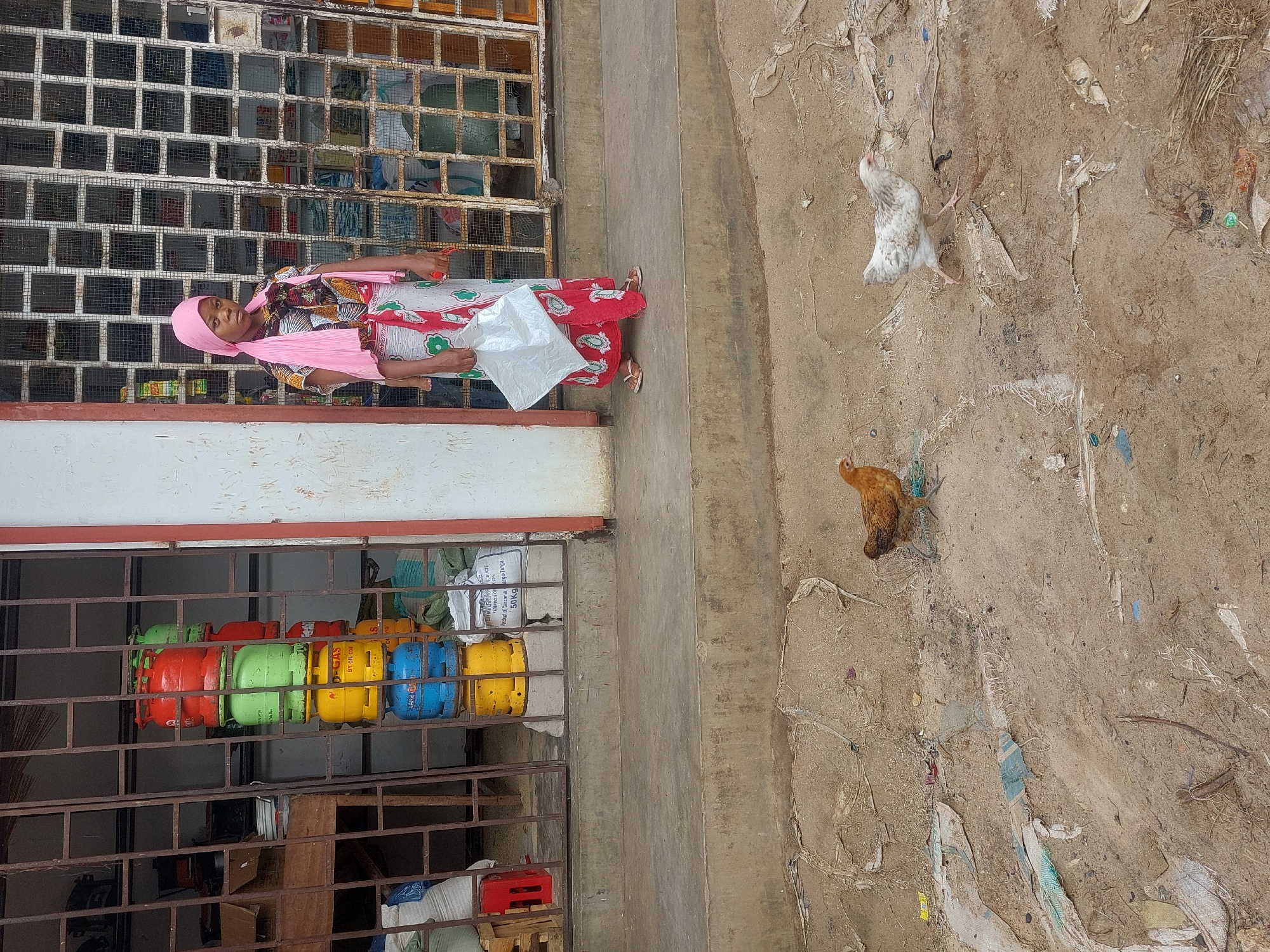 The image depicts an outdoor scene, likely a street in Dar es-Salaam, Tanzania, as indicated by the "Product of Tanzania" inscription on a 50kg gas cylinder visible in the lower left of the frame.
A woman stands in front of a building with a gated storefront. She is wearing a pink headscarf and shawl, a patterned dress, and carrying a white plastic bag. The storefront displays a variety of brightly colored gas cylinders (red, green, yellow, and blue) behind a metal grate.
To the right of the building, the ground appears to be dirt with scattered debris. Two chickens are visible in this area: one white and speckled, the other brown.
