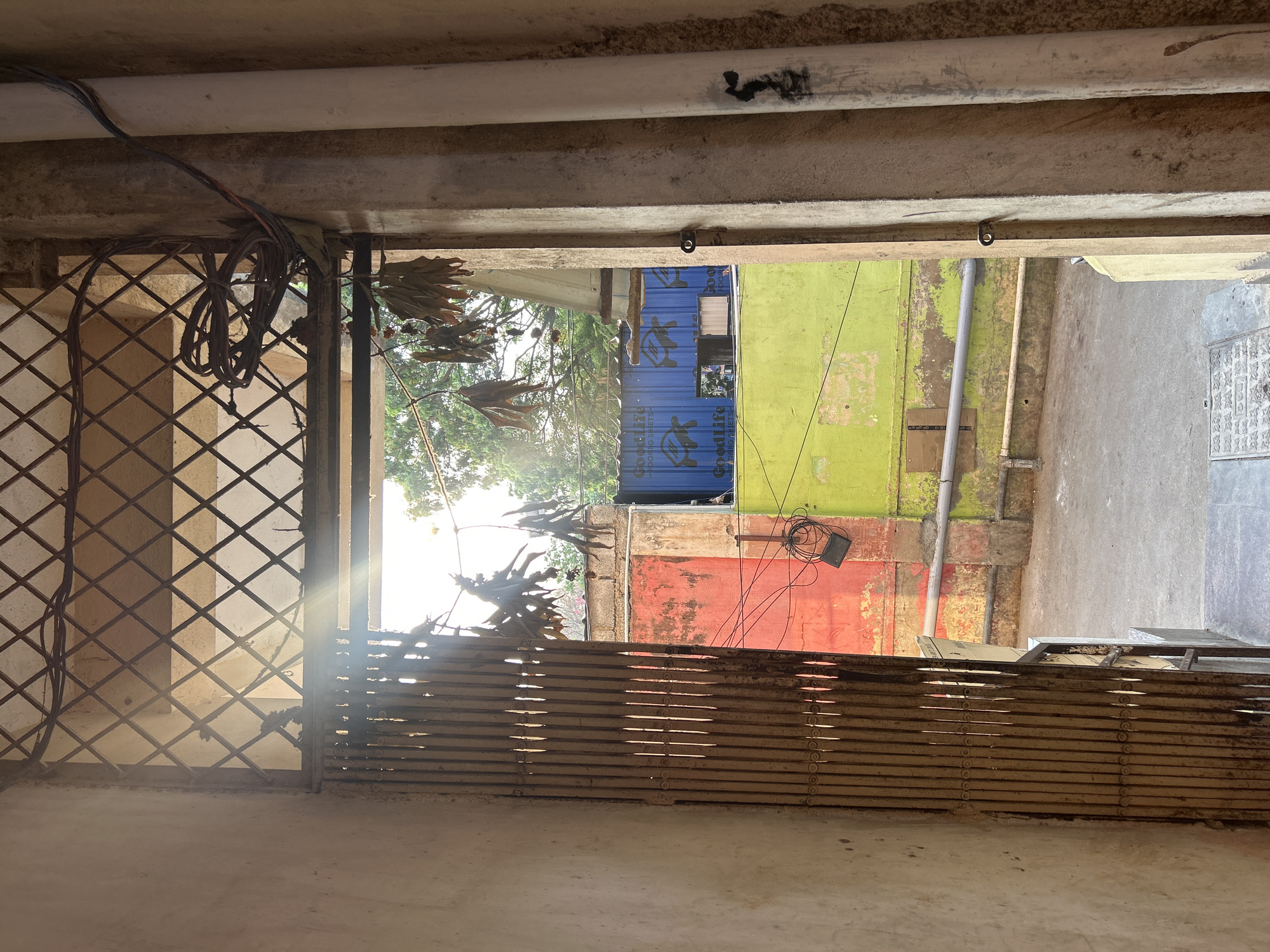 The image depicts a view from a building or structure in Puttaparthi, India, looking outward through a barred window or gate. The foreground features a metallic lattice gate on the left and a horizontal slat-like gate below. Wires and dried leaves are visible near the gate. Beyond the gate, the view opens to a narrow space between buildings. Walls in the background are painted in varying colors: red, green, and blue. A container-like structure with the word "Goodlife" is visible between the walls. An electrical box and wires are attached to the red wall. The environment appears to be urban and somewhat weathered. The light is bright, creating shadows and highlighting the textures of the walls and gates. The scene shows no people.