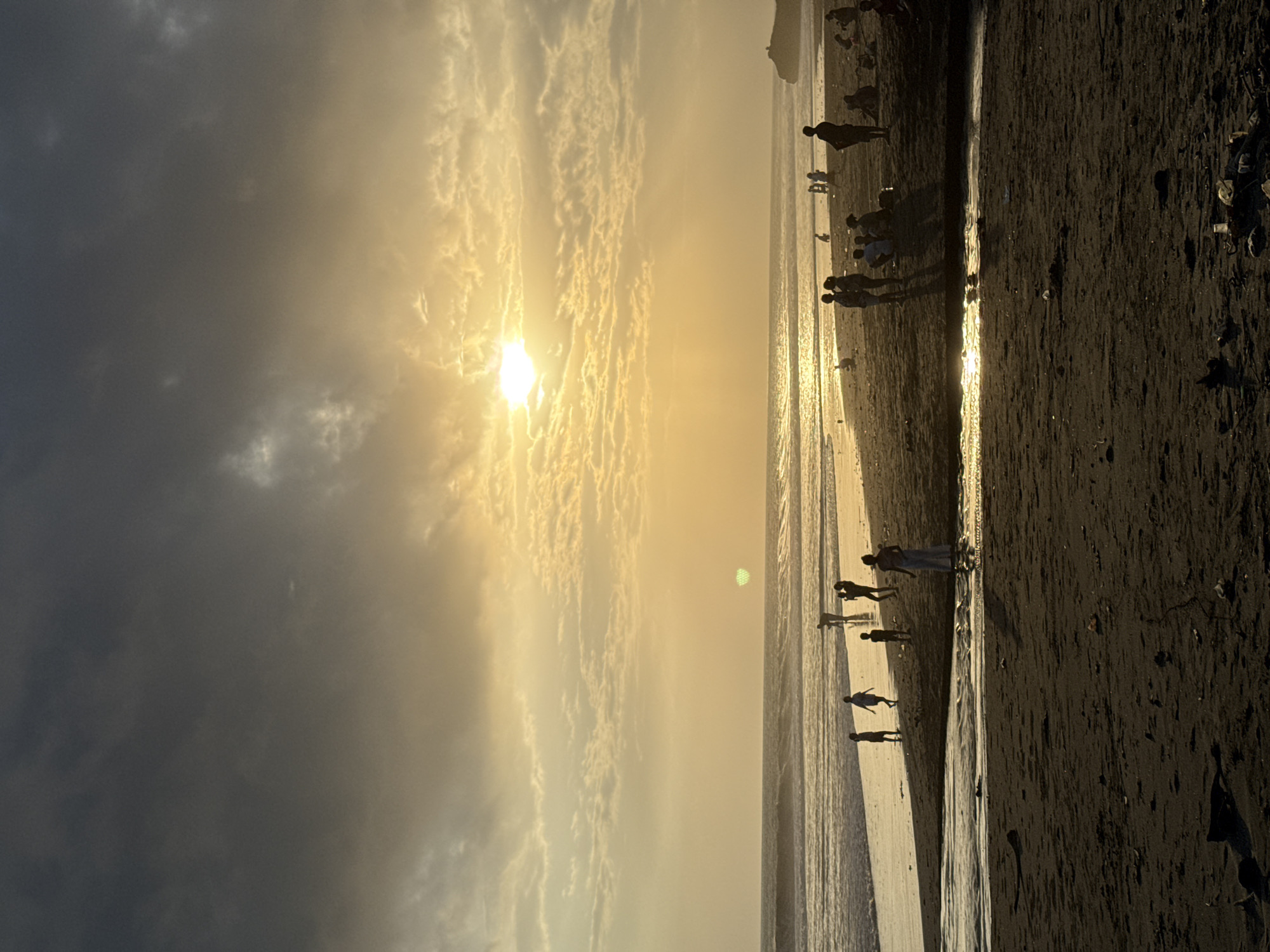 This image shows a beach scene at sunset in an unidentified location. The sun is positioned in the upper-center of the frame, obscured by thin clouds. The sky to the left of the sun is filled with darker, heavier cloud cover. To the right, the shoreline is visible, with dark sand and a shallow water line reflecting the sunlight. Silhouetted figures of people are seen walking along the beach at varying distances from the camera. The foreground shows close-up details of the dark sand with some debris scattered along the shore.