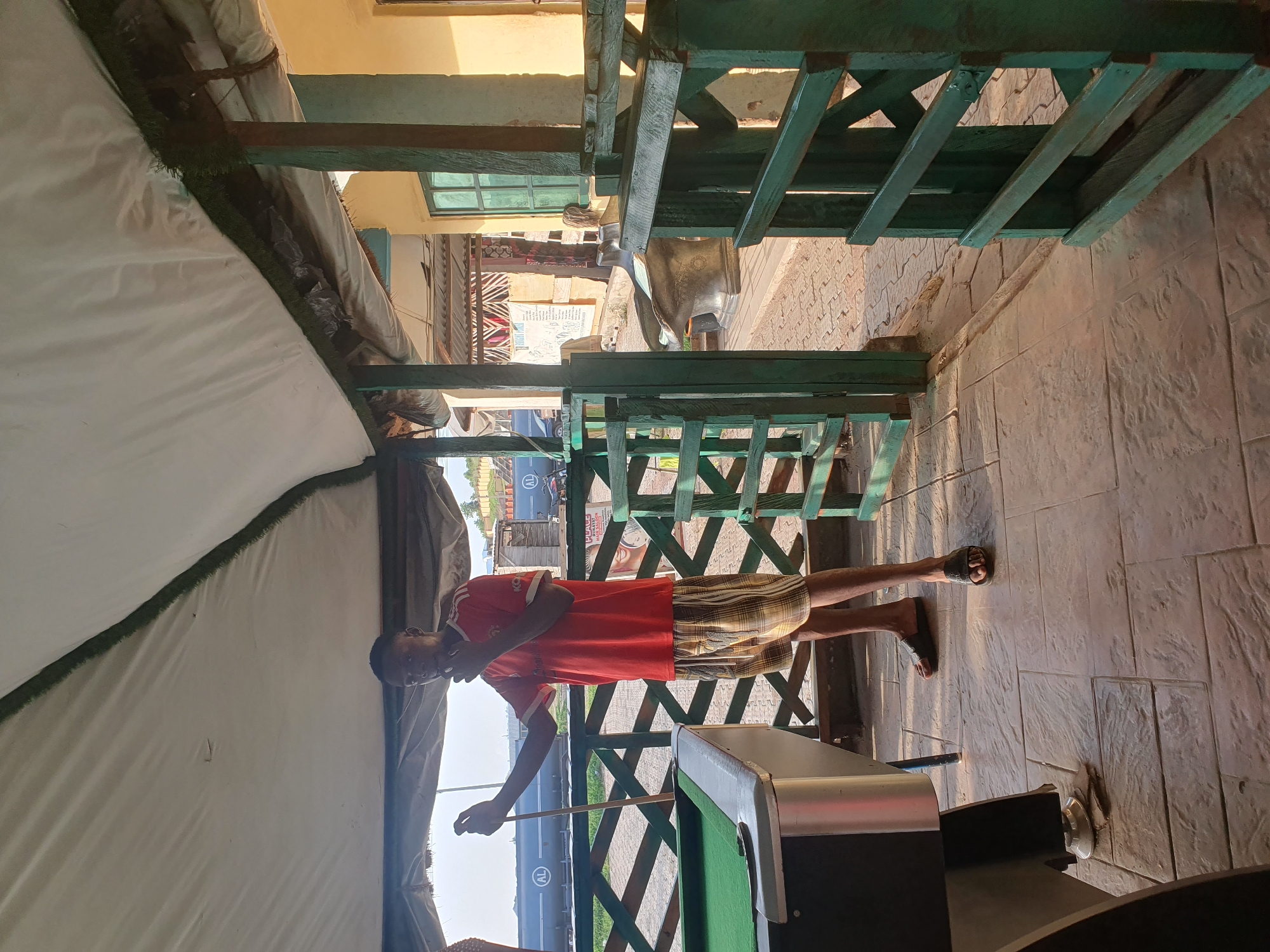 A young Black male, appearing to be in his late teens or early twenties, stands on a paved outdoor patio near a pool table. He is wearing a red t-shirt with white stripes on the sleeves and plaid shorts. He holds a pool cue in his right hand, resting it on the pool table. His left hand is raised to his mouth, as if in thought. The pool table has a green felt surface. Behind the man is a wooden railing and fence, painted green, separating the patio from an area with large tanks or containers. The area is covered by a white canvas awning supported by wooden posts. In the background, a building with yellow walls and green-framed windows is visible. The ground is paved with textured concrete tiles. The scene appears to be an outdoor recreational area, potentially a bar or a gathering spot, located in Lokoja, Nigeria.