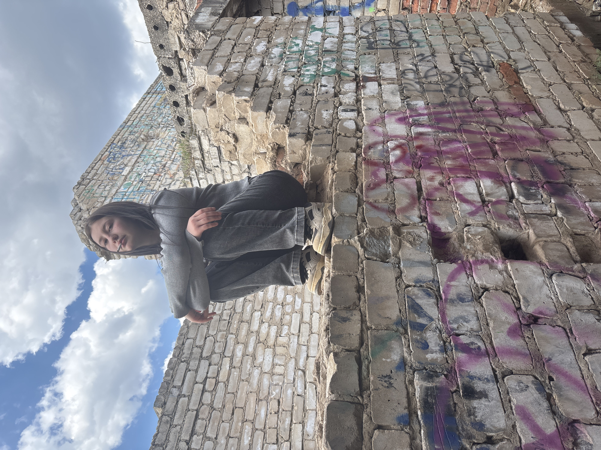 A young woman squats precariously atop a dilapidated brick structure, possibly the remnants of a building in Фоки, Russia. The structure is covered in graffiti of various colors. The woman is wearing a grey sweater and dark jeans, and appears to be smoking. The sky above is partially cloudy.