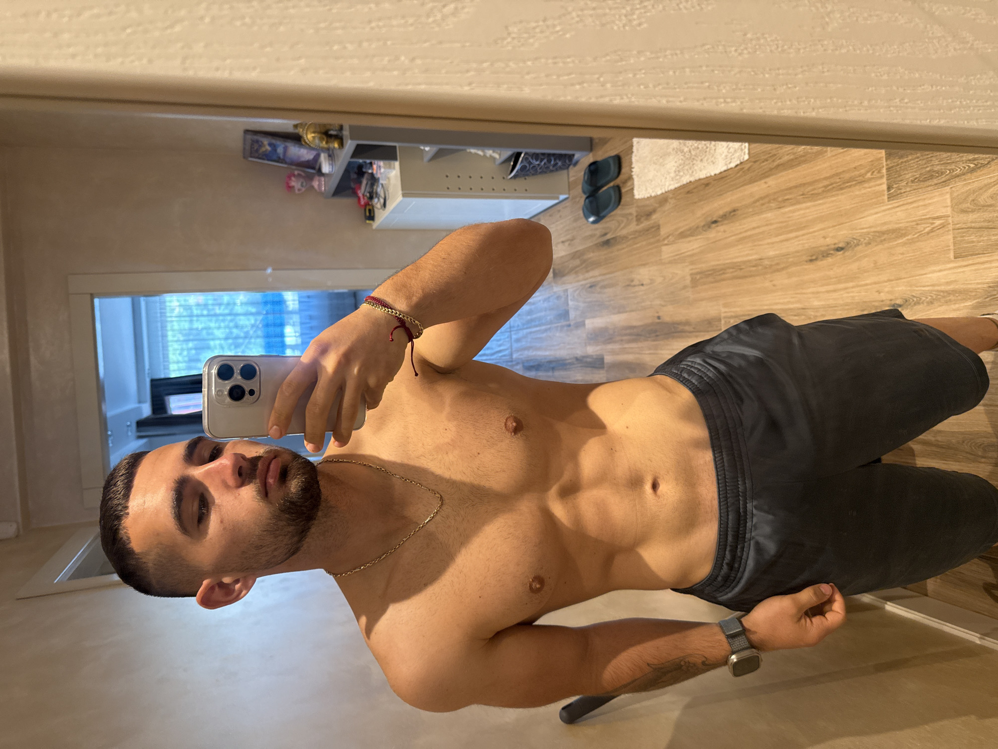 A young adult male is captured in a mirror selfie, standing shirtless and wearing dark athletic shorts. He holds a smartphone with a multi-lens camera in his left hand, which is visible in the reflection. He has short dark hair, a beard, a gold chain around his neck, and a red and gold bracelet on his left wrist. A smartwatch is worn on his right wrist.
The setting appears to be an indoor room, likely a residence in Gabrovo, Bulgaria, as reflected in the mirror. The background shows a light-colored, possibly textured wall. A window with white blinds offers a view of green foliage outside, suggesting daytime. The floor is made of light-colored wooden planks. Other reflected objects include a small white rug, a dark shelving unit with various items, and a pair of dark slip-on shoes on the floor. The man's expression is neutral as he looks at his phone while taking the picture.