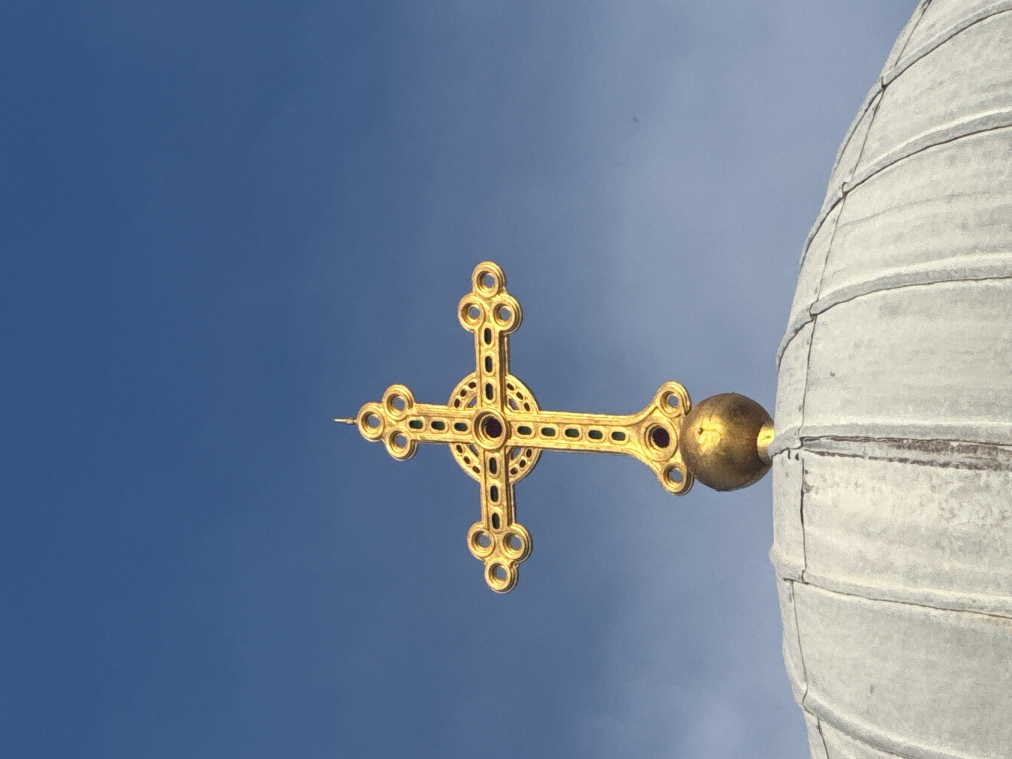 The image captures a close-up, low-angle view of a golden, ornate cross atop a dome, set against a clear blue sky.
**Subjects:** The primary subject is a radiant golden cross, elaborately decorated with circular patterns at the ends of its arms and intricate designs along its main beams. Small, dark, possibly gem-like inlays are visible within the golden framework of the cross. The cross is attached to a spherical golden finial, which then connects to a large, curved dome structure made of light-colored, possibly metal or stone, panels. The dome exhibits distinct horizontal banding and vertical seams, suggesting its construction. The background is a vibrant, cloudless blue sky, showing a slight gradient from a deeper blue on the left to a lighter, almost grayish-blue on the right.
**Activity or event taking place:** No specific activity or event is depicted; the image is a static architectural detail.
**Notable details:** The time of day appears to be sunny daytime, given the bright illumination of the golden cross and the clear sky. The weather is fair and cloudless. The ornate cross and dome are characteristic features of a religious building, likely a church or cathedral, situated in Sofia, Bulgaria, as per the location context. The photograph is taken from below, looking up, and is oriented so the cross appears horizontally across the frame rather than vertically, suggesting the camera was tilted. There are no people visible.
**Visible text:** No text is visible in the image.