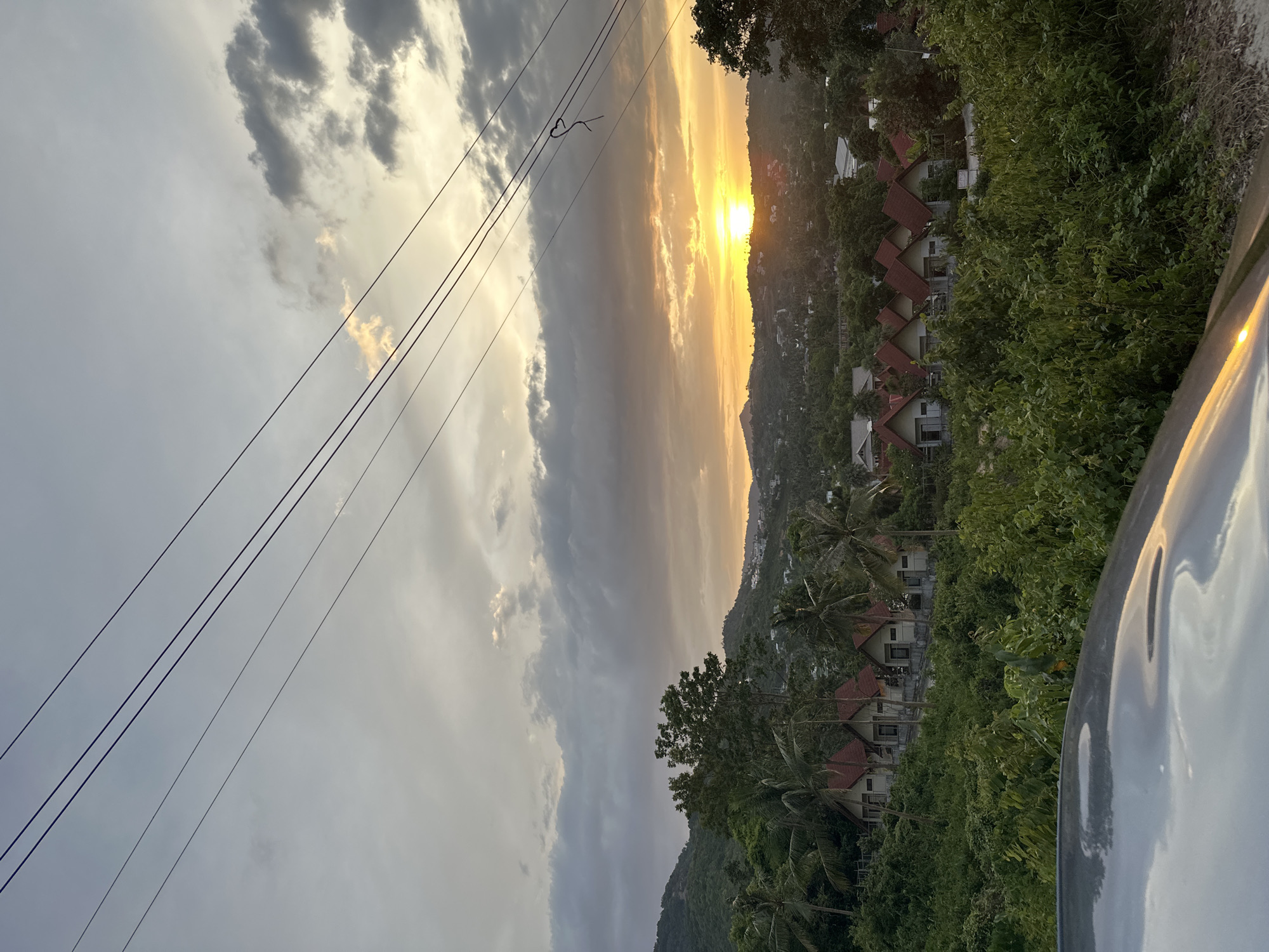 The image depicts a sunset over a lush, hilly landscape in the Ko Samui District of Thailand. In the foreground, a reflective surface, possibly the hood of a vehicle, occupies the bottom of the frame, showing a distorted reflection of the sky. Above this, a dense area of green foliage and trees dominates, with several small, uniformly designed buildings with red roofs nestled on a higher slope. These buildings appear to be houses or bungalows.
Further in the background, rolling hills covered in vegetation recede into the distance. The sky is filled with a dramatic sunset, characterized by a bright orange sun low on the horizon, casting a warm glow. Clouds are scattered across the sky, some illuminated by the sun's rays, creating a colorful and textured atmosphere. Four dark electrical wires stretch diagonally across the upper portion of the image, from left to right, adding a linear element to the natural scenery. The overall impression is one of a tropical evening scene, possibly in a residential or resort area.