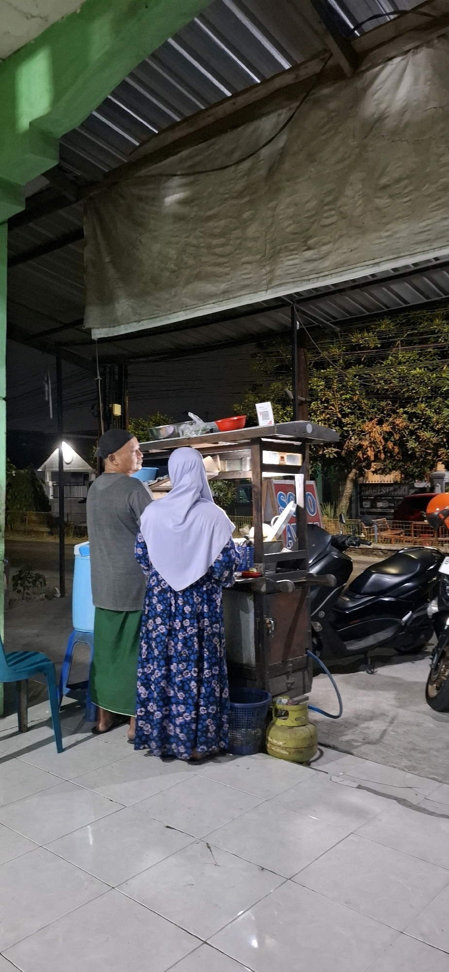 At night in Desa Gagaksipat, Indonesia, two people stand at a street food stall. The person on the left, wearing a dark cap, grey t-shirt, and green sarong, is looking to the right. The person on the right, wearing a light purple hijab and a blue floral dress, is facing the stall. The stall is wooden with a metal top, displaying food items and cooking equipment. A propane tank is visible on the ground next to the stall. In the background, there are parked motorcycles and trees. The ground is tiled.