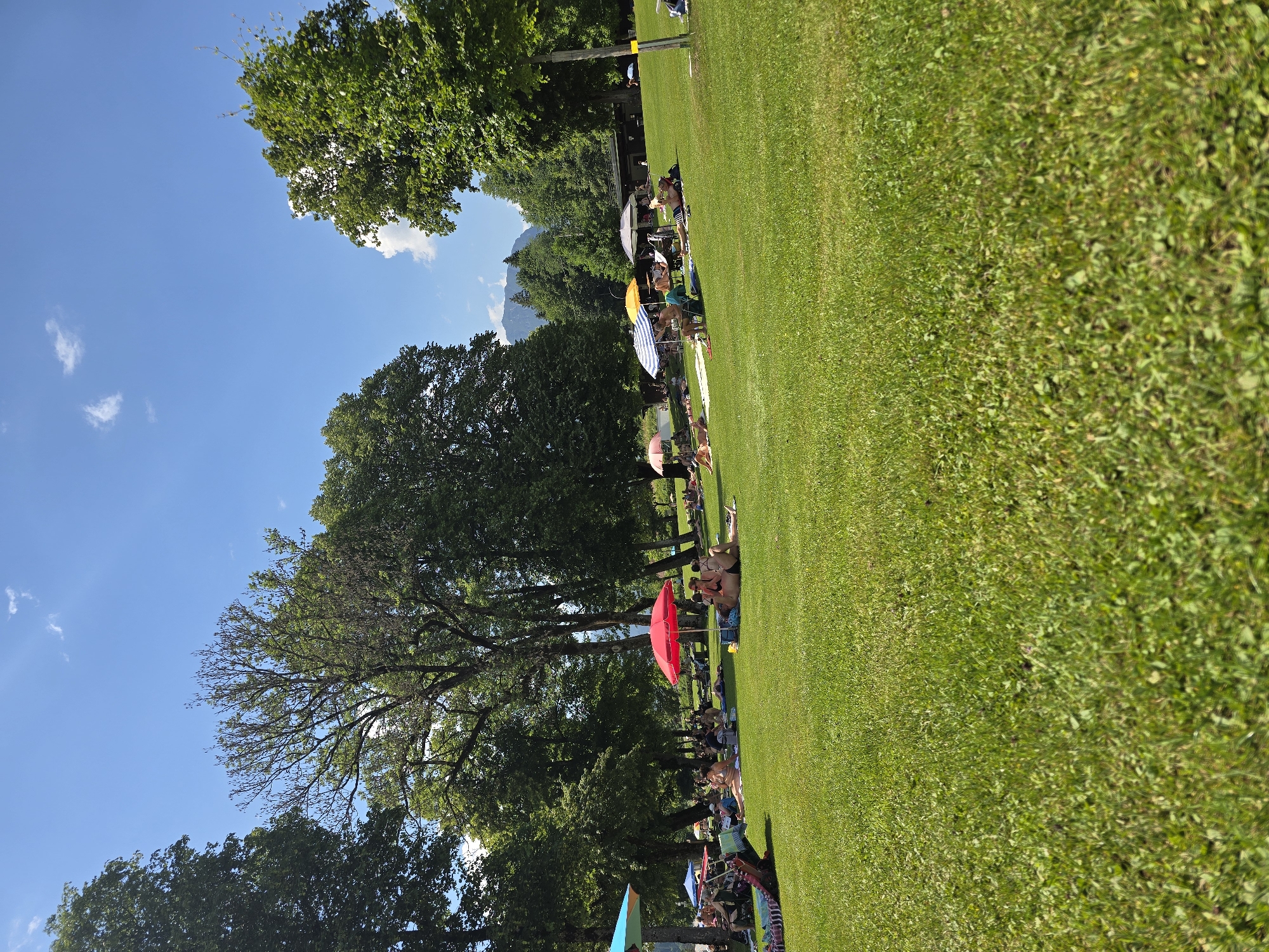 The image depicts a sunny day at a park or recreational area, likely in Garmisch-Partenkirchen, Germany, given the mountain visible in the background. The scene is dominated by lush green grass, with a line of people relaxing and sunbathing under the clear blue sky. Several umbrellas, some brightly colored like a prominent red one, are scattered across the lawn, providing shade for the sunbathers. Trees line the edge of the grassy area, with their leaves appearing vibrant green, suggesting it is summertime.
The people are casually dressed in swimwear and summer attire, indicating a leisure activity. Some are lying on towels or blankets, while others are seated. The overall atmosphere appears relaxed and cheerful, with individuals enjoying the warm weather and outdoor environment. The time of day is clearly daytime, with strong sunlight casting shadows and illuminating the scene. The presence of mountains in the distance further confirms a scenic, likely alpine, location. There is no visible text in the image.