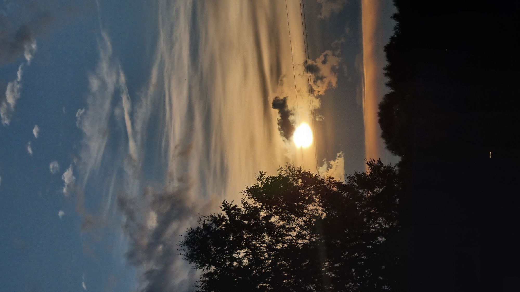 The image depicts a sunset scene in the city of Vysokoe, Russia. In the foreground, the silhouette of a deciduous tree is visible on the left, its branches stark against the twilight sky. The sun, a bright, luminous orb, is partially obscured by dark clouds, casting a warm, orange glow across the horizon. Wispy clouds stretch across the sky, painted in hues of blue, orange, and gray. A line of dark trees forms a silhouette along the distant horizon. The lower portion of the image is a deep, dark silhouette, obscuring details of the ground and any potential human presence. A faint, small, rectangular light source is visible in the very bottom center of the image. The overall atmosphere is serene and natural.