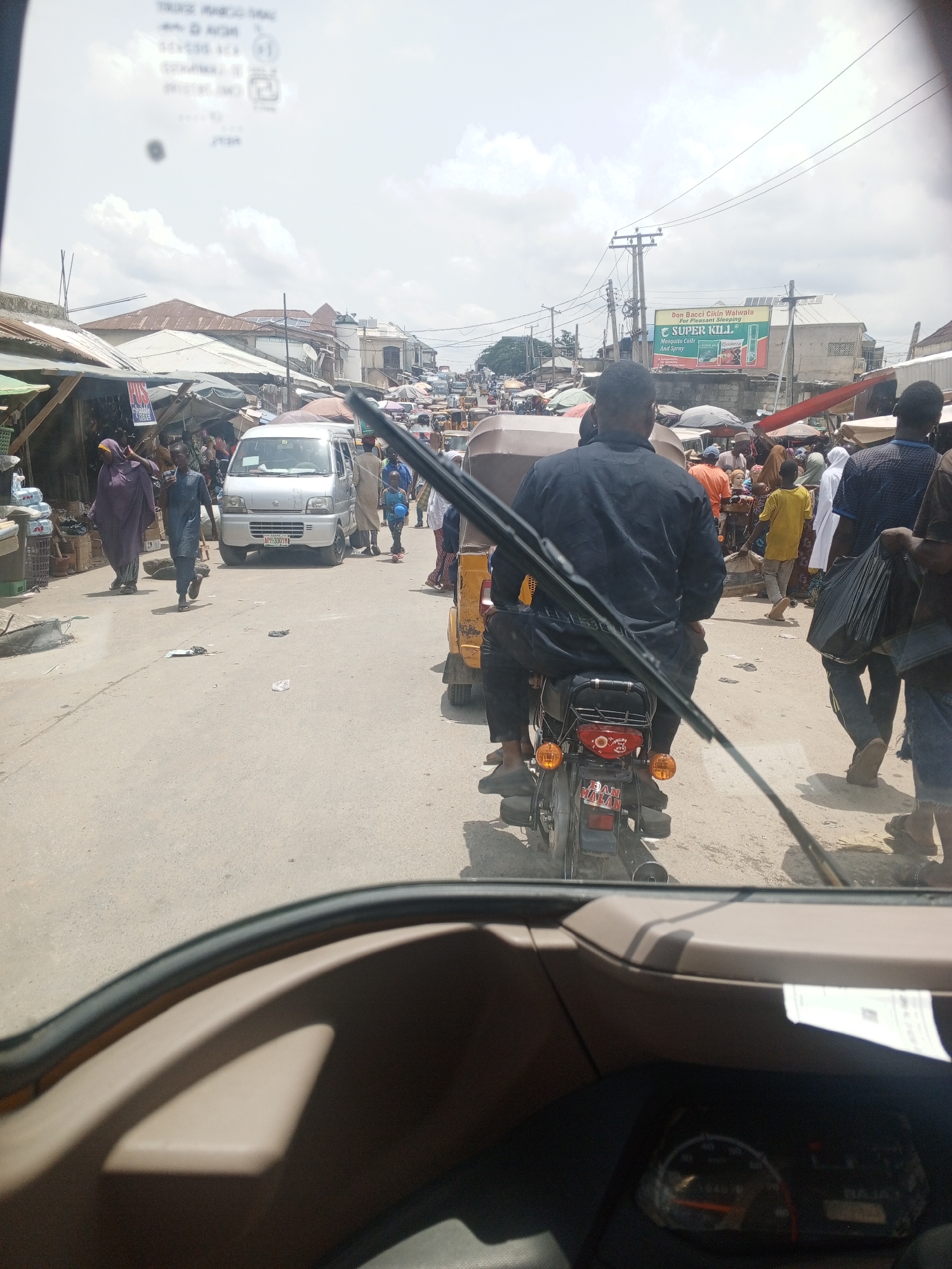The image depicts a bustling street scene, likely a market area, in an urban environment. The view is taken from inside a vehicle, focusing on the road ahead. A motorcycle with two riders is directly in front, partially obscuring a white van and numerous pedestrians. The scene is filled with people engaged in various activities, suggesting a lively marketplace. Buildings and makeshift stalls line the street, and a large billboard advertising mosquito coils and spray is visible in the background. The presence of power lines overhead further indicates an urban setting.