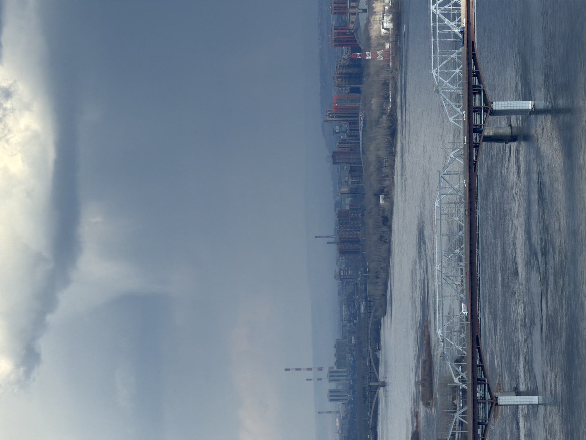 The image presents a wide, panoramic view of a city along a river under an overcast sky. The scene is dominated by a broad, dark river occupying the foreground and mid-ground on the right side of the frame. Spanning this river is a prominent, multi-span truss bridge, likely for railway traffic, characterized by its intricate metal framework in shades of grey and rust brown, supported by pillars in the water.
Along the far bank of the river, a sprawling city or industrial area stretches into the distance under a hazy sky. Numerous buildings, including what appear to be factories and other industrial structures, form a dense skyline. Several tall, slender smokestacks, some featuring distinctive red and white bands, rise above the other buildings, suggesting heavy industry. The riverbanks are lined with bare trees, indicating a season like late winter or early spring.
The sky on the left side of the frame is heavily clouded with thick, white and grey formations, while further right, over the city and river, the sky appears a more uniform, muted grey, hinting at a dull, possibly misty, daytime. No direct sunlight is visible, and the overall lighting is diffuse.
There is no discernible human activity or specific events taking place in the image; it captures a static landscape. No visible text can be clearly identified on any of the structures. Given the location context, the scene likely depicts the Yenisei River flowing through Krasnoyarsk, Russia, with its industrial complexes and infrastructure. The atmosphere conveyed is rather industrial and somewhat stark due to the weather and urban landscape.