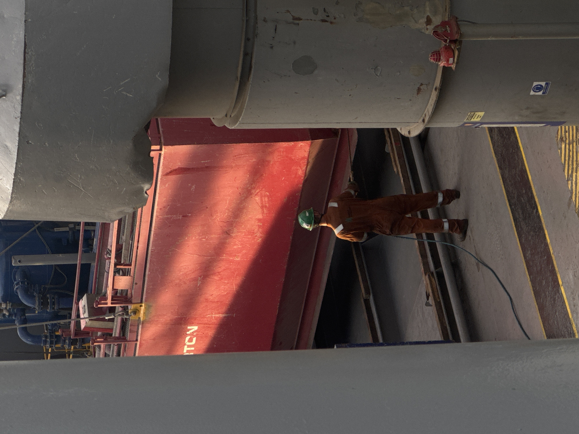 The image shows a person wearing an orange jumpsuit and a green hard hat, holding a coiled blue hose. The person is standing on a gray surface marked with yellow lines, next to a large red structure. In the background, there is a blue industrial machine with pipes and valves. The scene appears to be on a dock or in an industrial area.