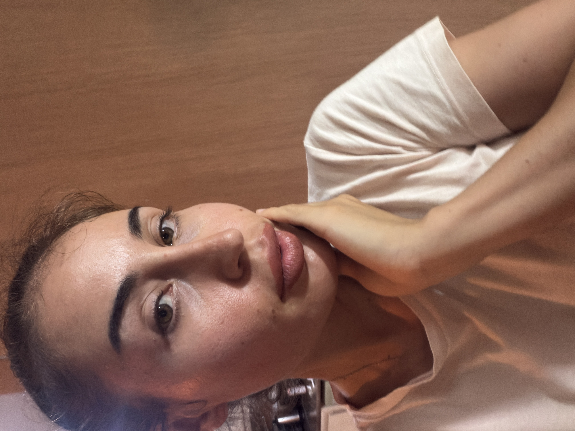A medium shot portrait of a woman with brown hair pulled back is captured in a dimly lit room with wooden-paneled walls. The woman is looking at the camera and has her right hand resting on her cheek. She is wearing a light-colored t-shirt. The lighting creates a warm glow on her skin, highlighting its texture. There is a hint of a necklace visible. The background is a plain wooden wall, suggesting an indoor setting. The image is devoid of any specific urban landmarks that would place it in Tersefanou, Cyprus.