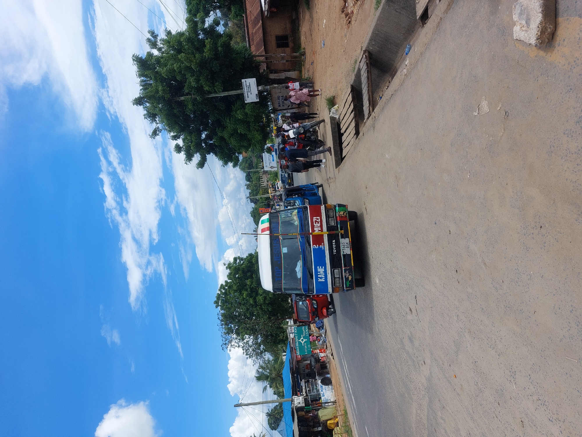 The image depicts a street scene in Dar es-Salaam, Tanzania. A "daladala" bus, a common form of public transport, is in the foreground. The bus is colorfully painted and has "KAME" and "MBEZI" written on it, along with "TOYOTA" and the license plate number "T665 AGK". Passengers are seen near the bus.
In the background, the image contains trees and buildings. Road signs indicate locations and distances: "Mbezi Mwisho 7 km", "Kimara", and "Tegeta 13 km". A street is visible.