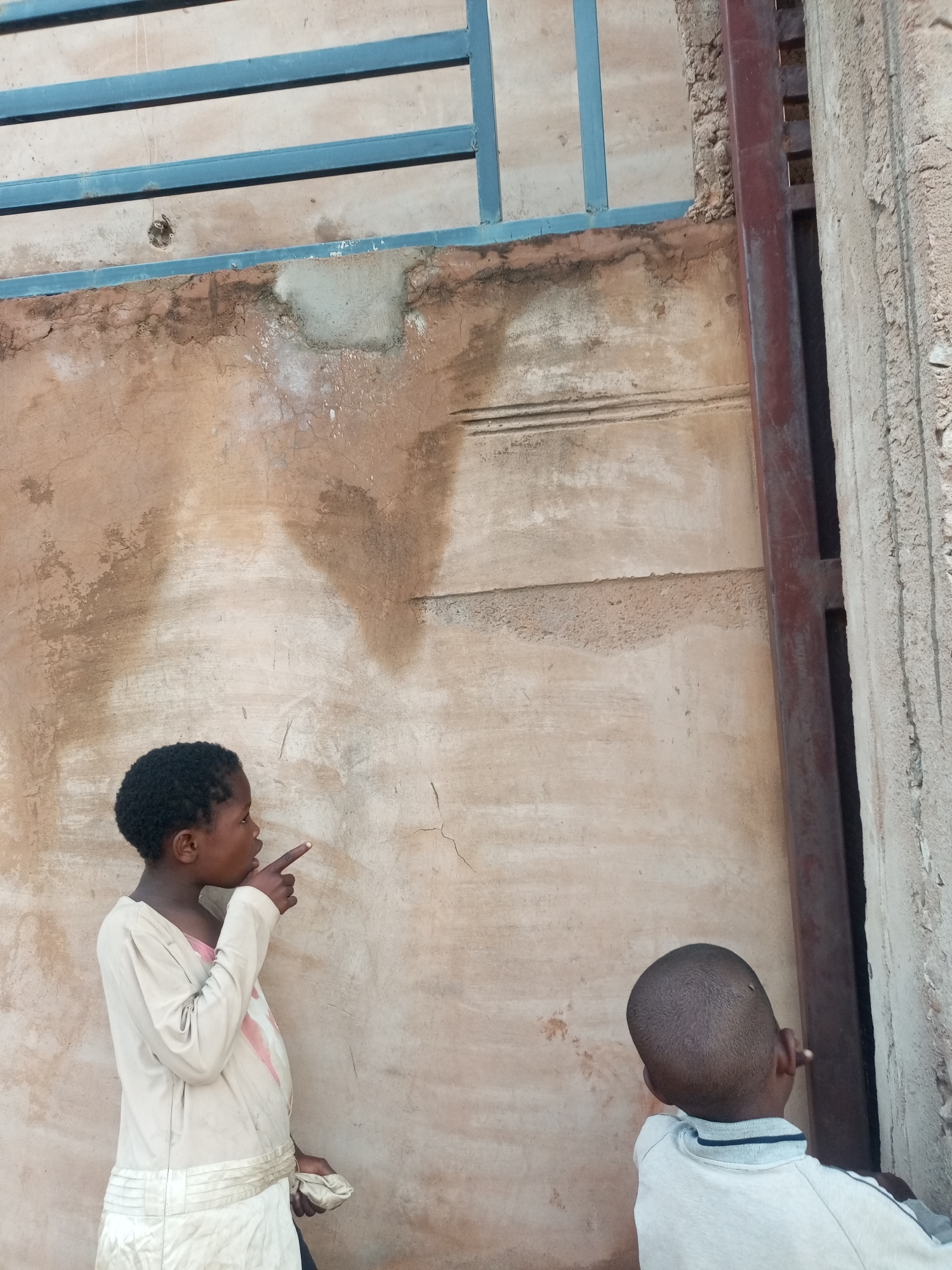 In Tempure, Nigeria, two young individuals stand near a weathered, ochre-colored wall. The individual on the left, facing right, wears a cream-colored tunic with light pink accents and holds a white cloth. They are pointing a finger toward the wall. The second individual, whose back is to the camera, wears a light gray collared shirt and appears to be looking at the same wall. Above the wall, a blue metal grating runs horizontally. A dark red metal beam runs vertically along the right edge of the image, adjacent to the wall. The wall itself shows signs of age, with areas of plaster damage and staining.