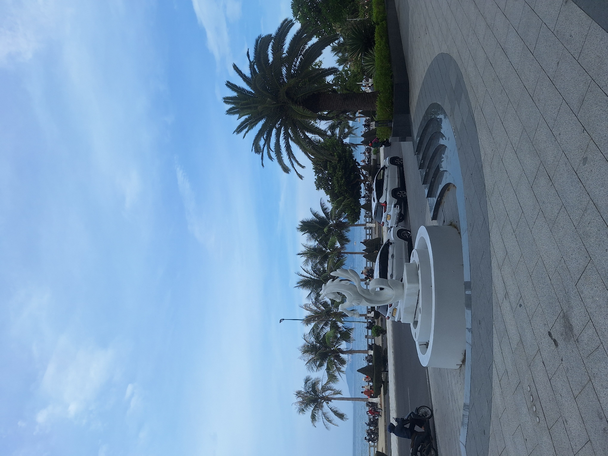 The image depicts a daytime scene along the coastline of Nha Trang, Vietnam. In the foreground, a large, circular stone structure, possibly a fountain base, is visible, with a white abstract sculpture resembling a stylized fish or sea creature situated on top. A wet, multi-tiered curved water feature is adjacent to the fountain. Paved walkways with stone tiles lead away from the fountain area. A road runs parallel to the coastline, with several white cars parked or driving along it, including a white SUV. A person on a dark-colored motorcycle is seen riding on the road in the left foreground.
Beyond the road, a row of palm trees lines the beachfront. The ocean stretches out to the horizon, with a calm sea and a distant hazy coastline. People are visible walking along the beachfront and near the palm trees. In the background, the sky is a pale blue with scattered white clouds. The overall atmosphere suggests a public area, likely a promenade or park area near the beach.
