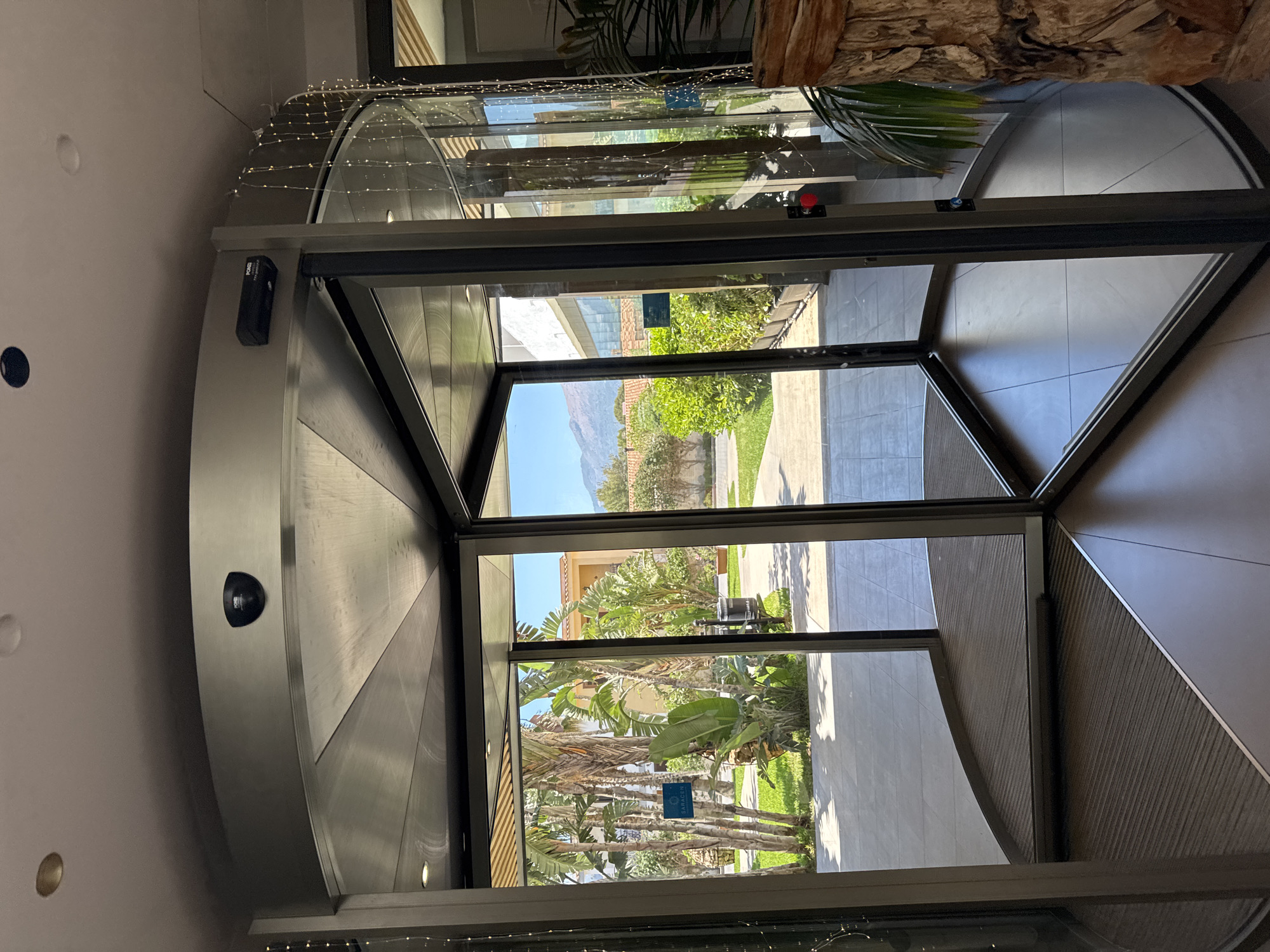The image shows a revolving door at the entrance of a building, likely a hotel or resort, in Isola delle Femmine, Italy. Through the glass panels of the door, a sunlit courtyard with palm trees, lush green foliage, and Mediterranean-style buildings is visible. Mountains can be seen in the background under a clear blue sky. Inside the entrance, the revolving door mechanism is a prominent feature, made of metal and glass. Small decorative lights are strung along the top edge of the door frame, adding a festive touch. There are no people visible in the image. The scene suggests a warm, pleasant climate, characteristic of the Italian coast.
