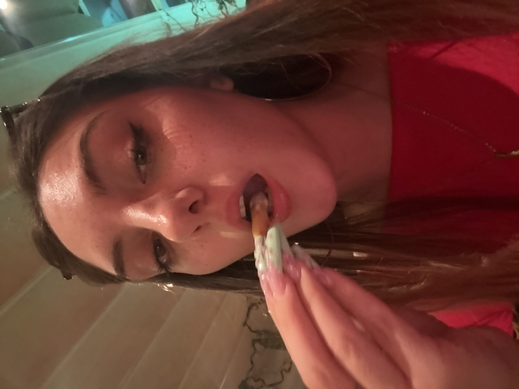 A young woman with light skin and long brown hair is holding a cigarette in her mouth and a finger-painted, green-stained cigarette in her hand. She is wearing a red shirt and large hoop earrings. The background is unclear, but it appears to be an indoor setting. There is no information available to confirm the location in the city of Sofia, Bulgaria.