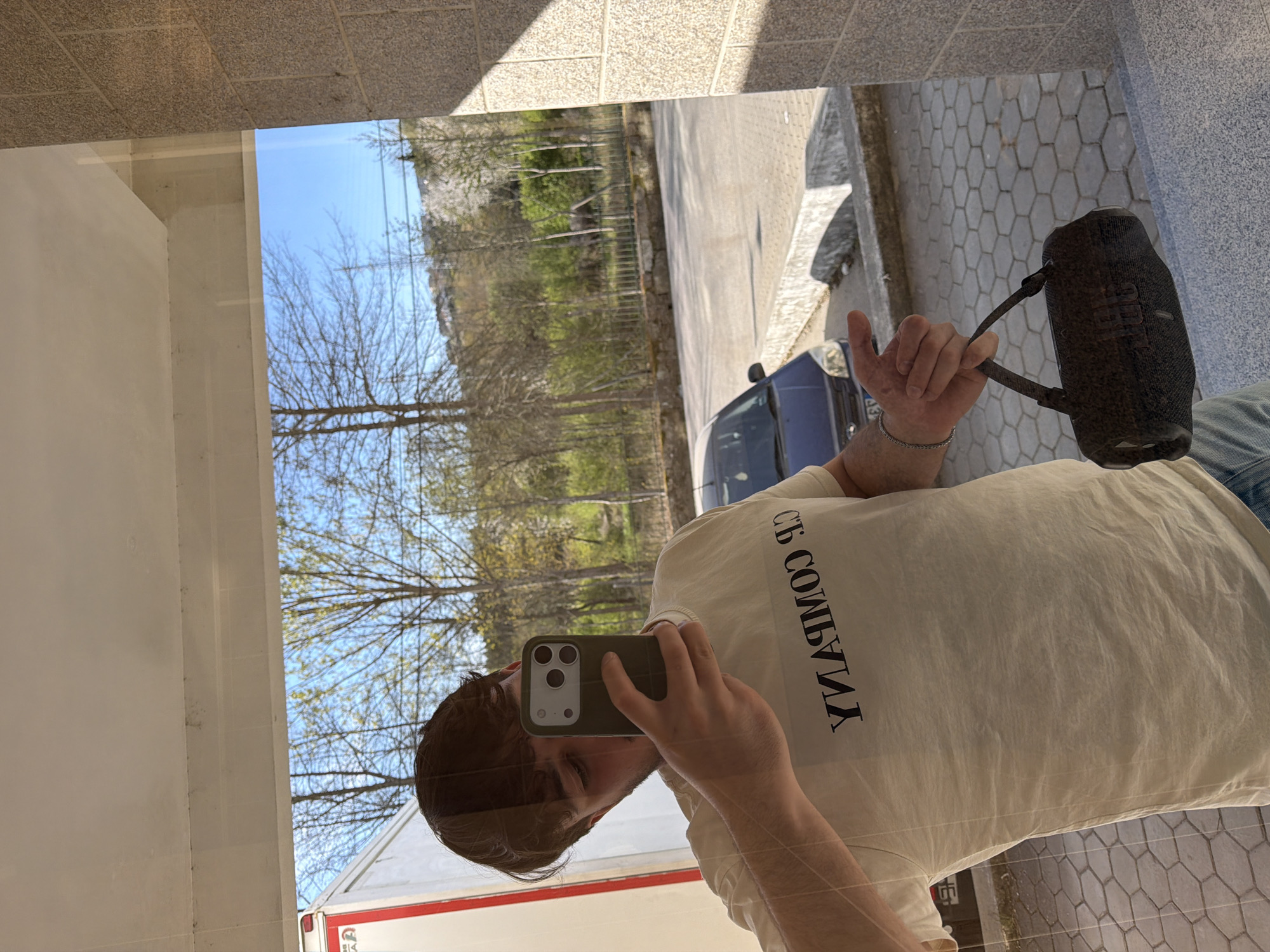 A man is reflected in a window while taking a selfie with a smartphone. The man is wearing a light-colored t-shirt with text printed on it. He is holding a black JBL speaker in his right hand. The reflection in the window shows a street scene, including trees, a blue car, and a white truck with red accents. The location appears to be in Vila Real, Portugal. The foreground shows tile flooring.