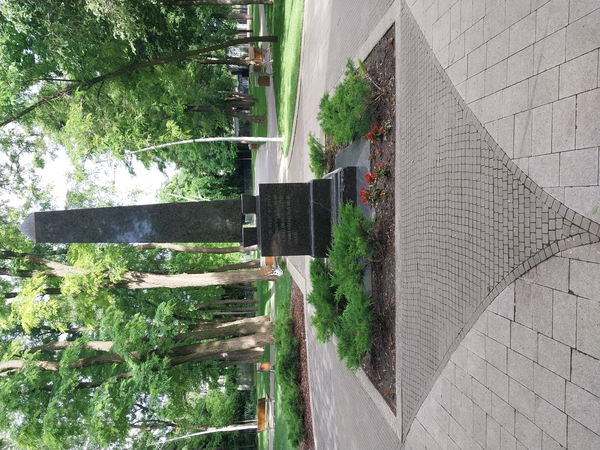 The image shows a war memorial in Dnipro, Ukraine. The memorial is an obelisk made of dark stone, with an inscription on its base. It is surrounded by a circular flower bed with green bushes and red flowers. The memorial is situated in a park with tall trees, green grass, and paved walkways. Benches are visible in the background. The time of day appears to be daytime, with natural lighting. There are no people visible in the image.