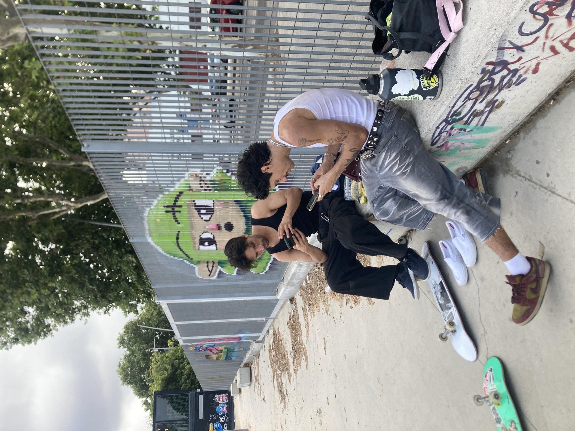 Two young men are sitting on a concrete ledge in front of a metal fence adorned with graffiti. Both individuals are looking at their smartphones. The man on the left wears a black tank top and dark, wide-legged pants. The man on the right wears a white tank top, grey jeans, and a studded belt. Skateboards and shoes are scattered on the ground around them. The background shows trees and some buildings. The scene appears to be an urban outdoor environment, possibly a skate park or an area adjacent to one. There is no specific indication in the image to place the location in Badalona, Spain.