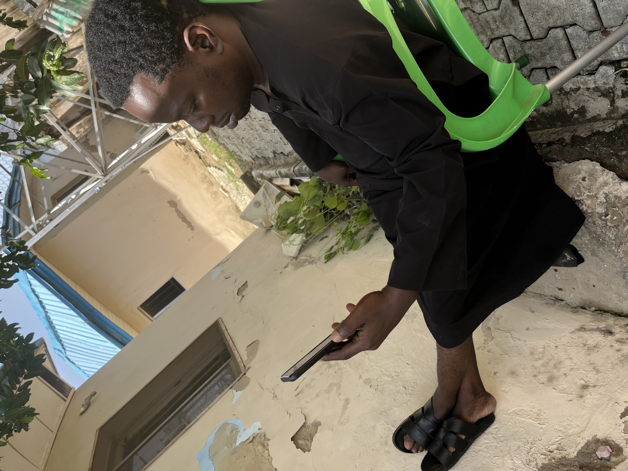 A medium shot captures a man sitting outdoors, wearing a black traditional outfit and black sandals, and holding a smartphone in his right hand. He is seated on a bright green plastic chair, positioned on a paved stone surface. The man's gaze is directed downwards at the phone.
The background reveals a building with a cream-colored textured wall, showing signs of wear and tear. A window with a metal grate is visible on the wall. Above, a light blue corrugated metal roof is visible, extending from the building. To the right, a metal structure, possibly part of a larger construction or support system, is present. Lush green leaves from an overhead tree cast shadows on the scene. The lighting suggests it is daytime. The location is identified as Galadimawa Village, Nigeria.