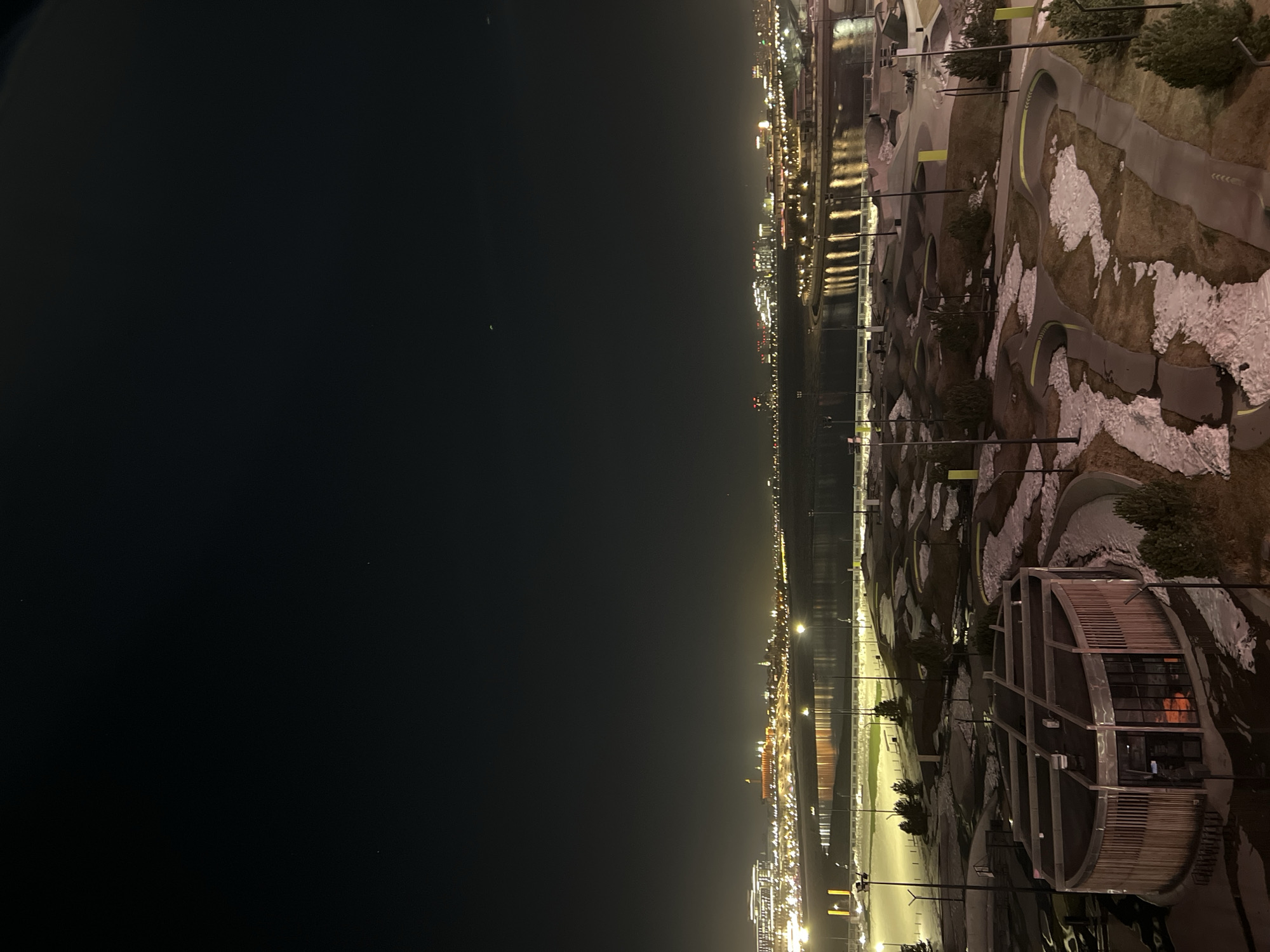 The image shows a night view of Kazan, Russia. The foreground features a modern park with curved paths and landscaping, including a distinctive circular building with a slatted facade. Light snow patches are visible on the ground. A body of water reflects city lights, separating the park from the illuminated cityscape in the distance. Streetlights are interspersed along the park's pathways and the waterfront. The sky is dark and largely empty.