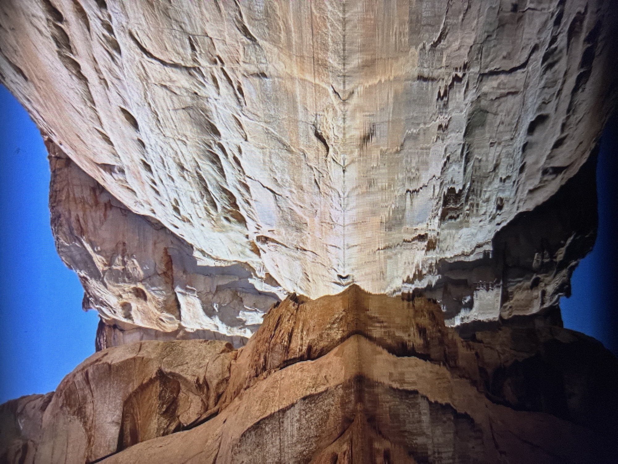 The media file displays a photograph of impressive natural rock formations, captured from a low-angle perspective looking upwards towards a massive overhead rock structure. The dominant subject is a vast, light beige to off-white rock ceiling or overhang, characterized by intricate textures, deep grooves, and patterns consistent with natural erosion and weathering. These textures create a series of ripples and channels across its surface.
Below this large overhang, darker, reddish-brown rock formations are visible, forming the lower part of the scene. These rocks exhibit more angular shapes and distinct layers, suggesting a different type of geological structure or a different erosion pattern. Patches of bright blue sky are visible on the left and right edges, indicating an outdoor setting with a clear sky.
No people or discernible activity are present in the image. The strong contrast between light and shadow suggests bright daylight, implying a clear weather condition during the time of capture. The natural formations evoke a sense of grandeur and timelessness. There is no visible text.
The image itself appears to be a photograph taken of a screen, evidenced by the visible pixel grid, particularly noticeable in the cropped hint images. The location context provided is Asenovgrad, Bulgaria, suggesting these geological features are found in that region.